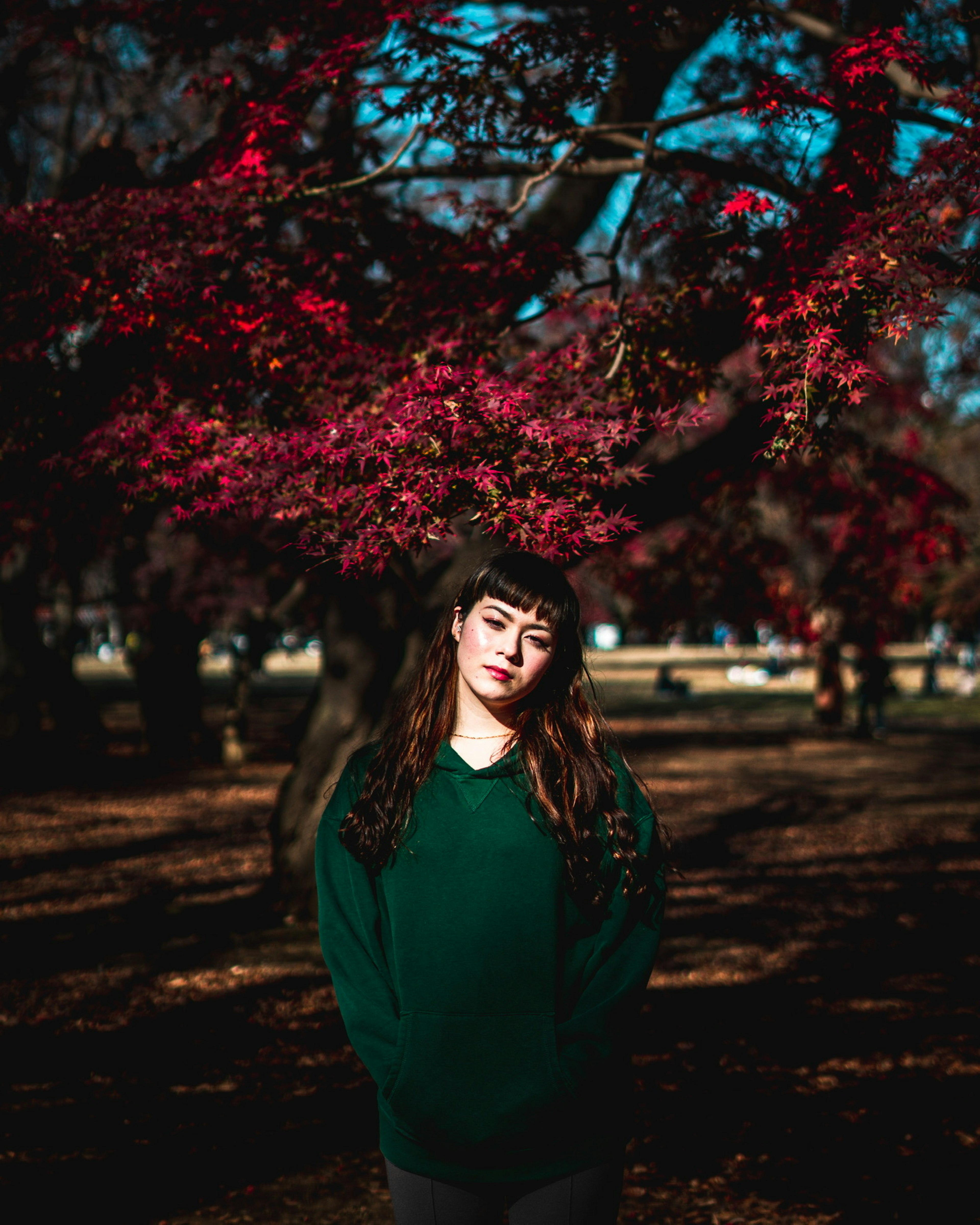 Perempuan dengan sweater hijau berdiri di bawah pohon dengan daun merah