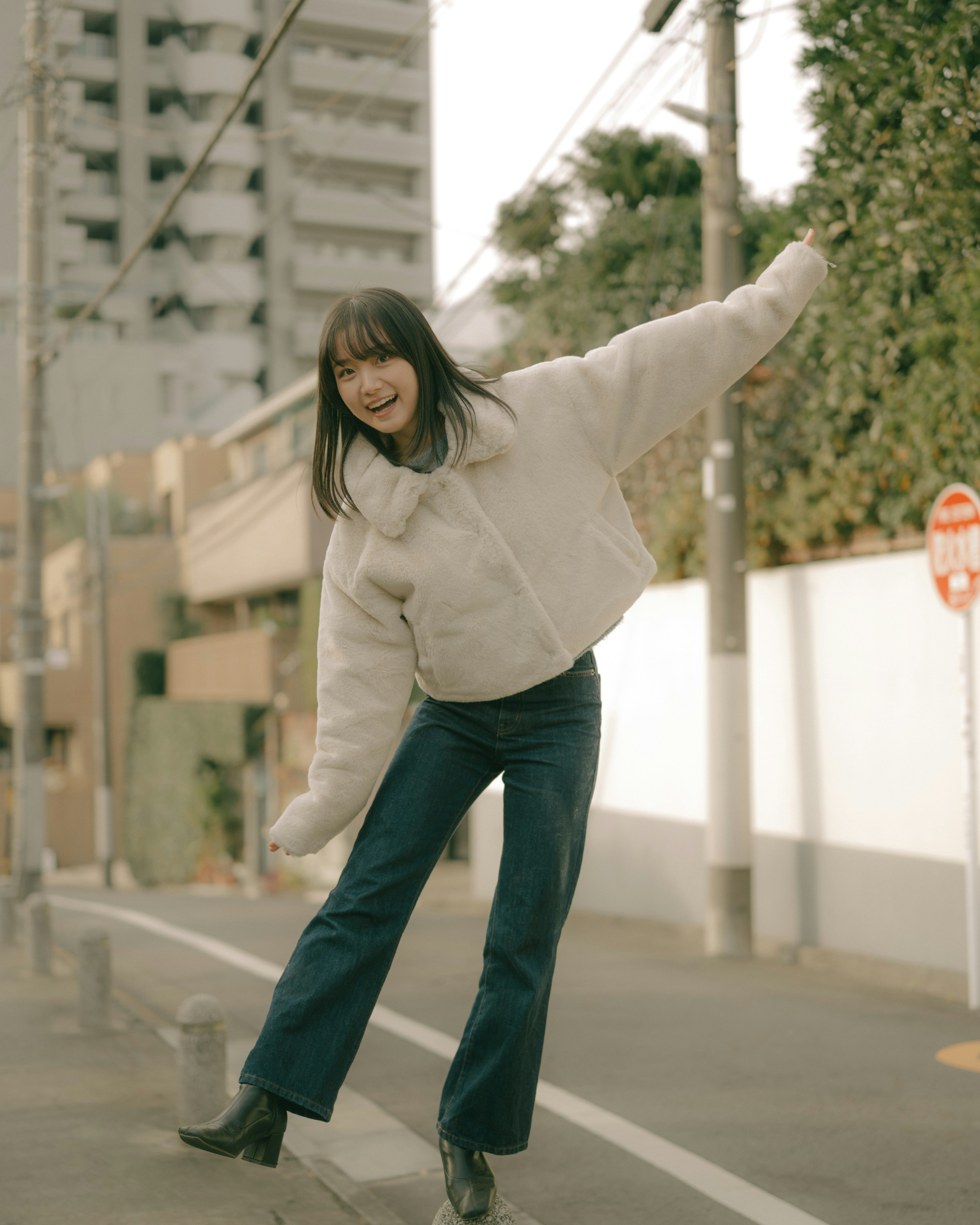 A woman joyfully balancing on a street, wearing a white jacket and denim, buildings and greenery in the background