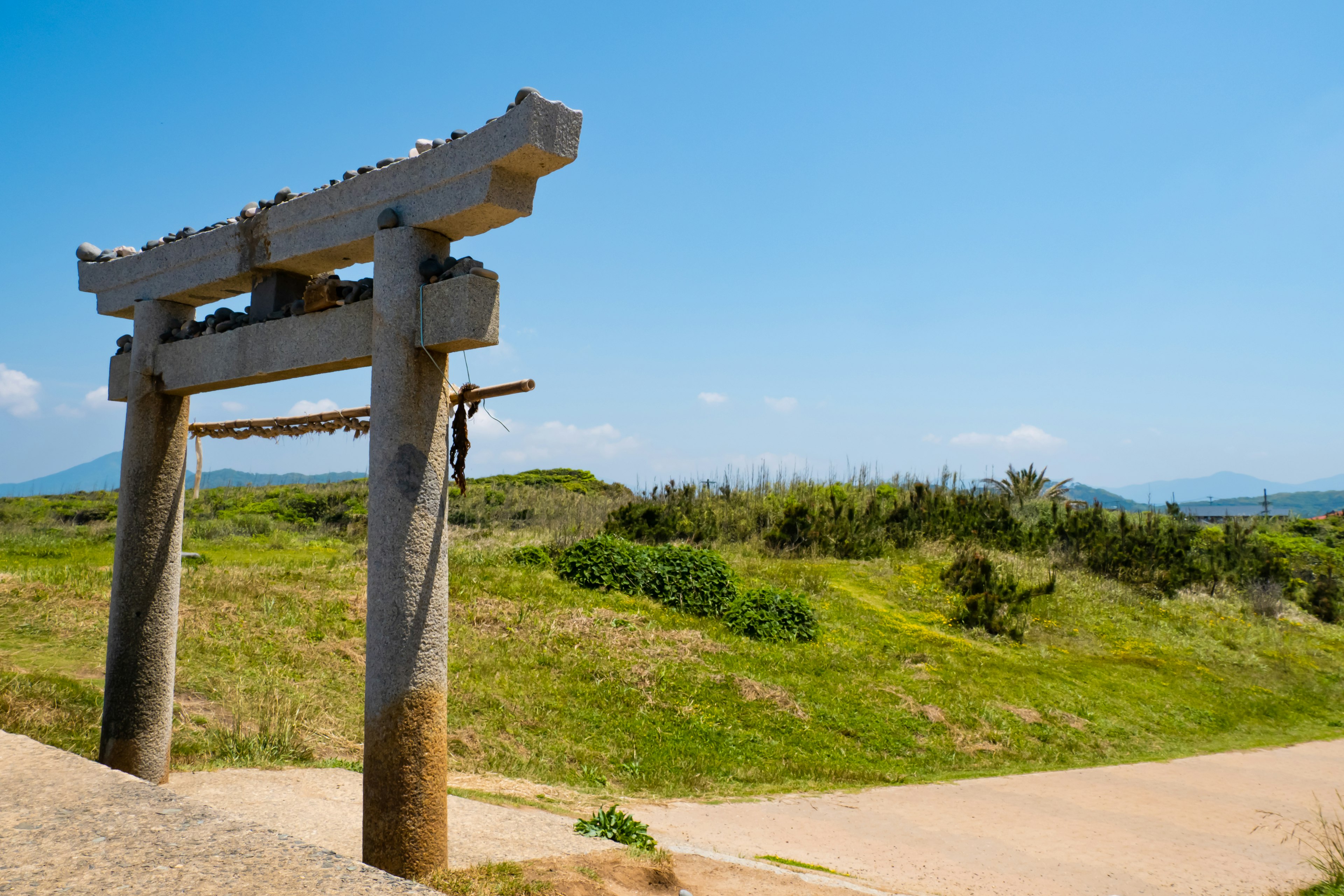 Gerbang torii batu di bawah langit biru cerah dengan bukit hijau