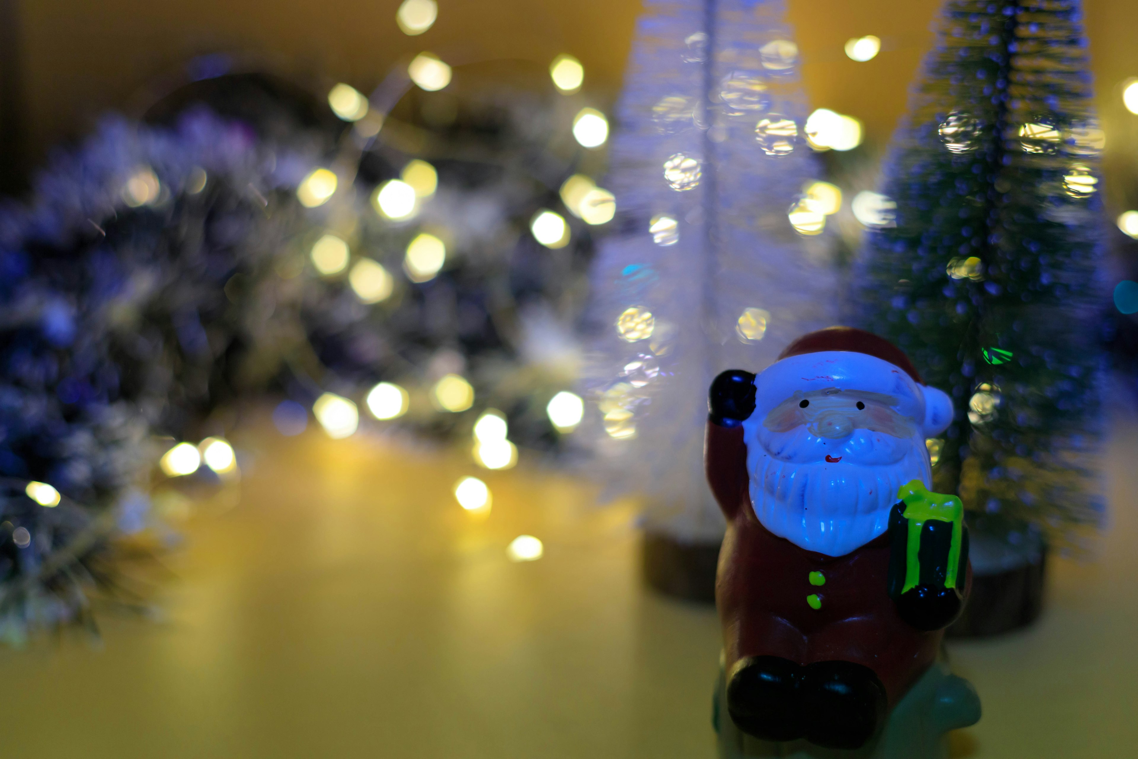 Santa Claus figurine standing in front of small Christmas trees with blurred light bokeh in the background