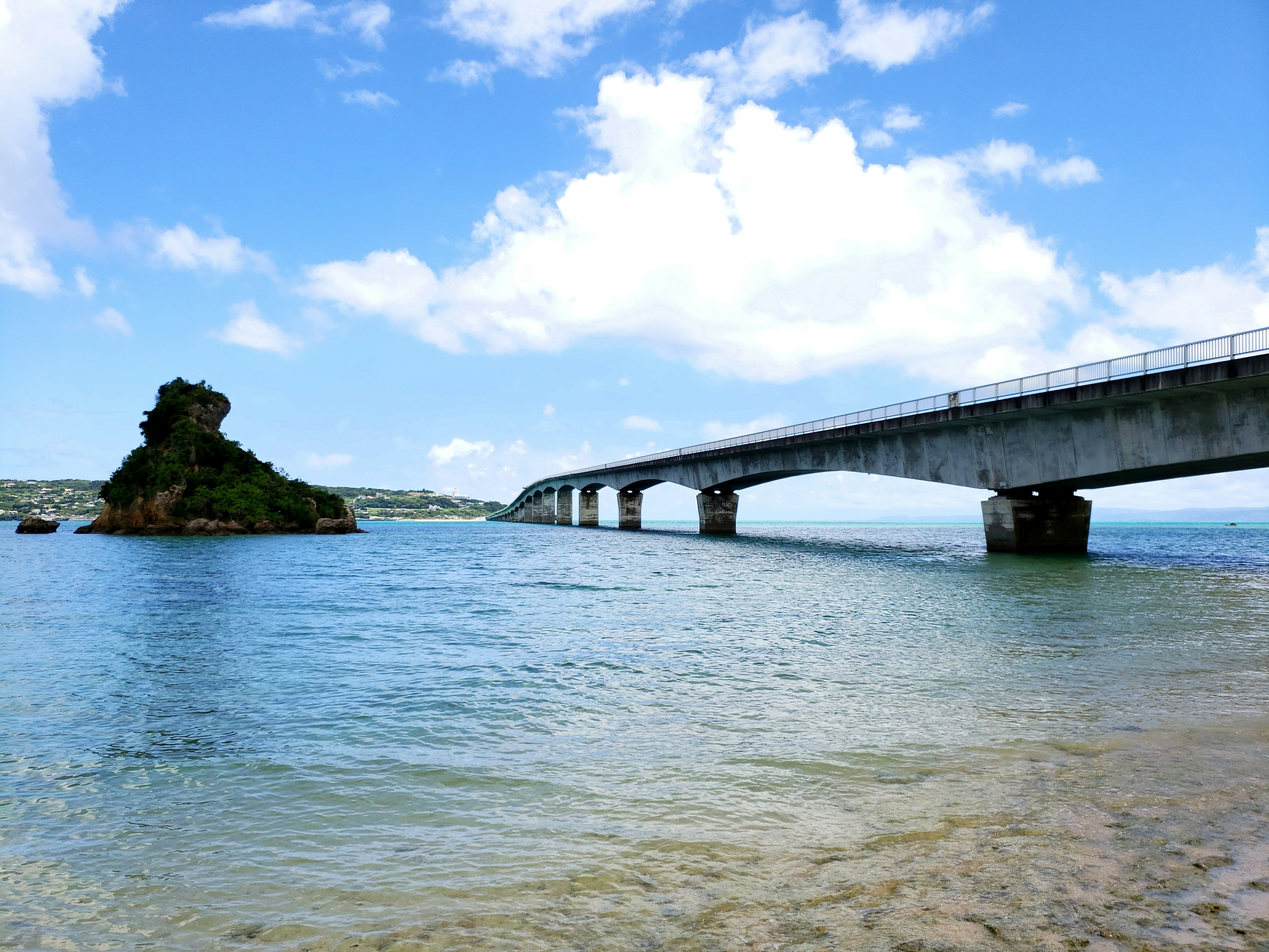 Pemandangan indah air tenang di bawah langit biru dengan awan putih sebuah jembatan melintasi air dan sebuah pulau berbatu di dekatnya