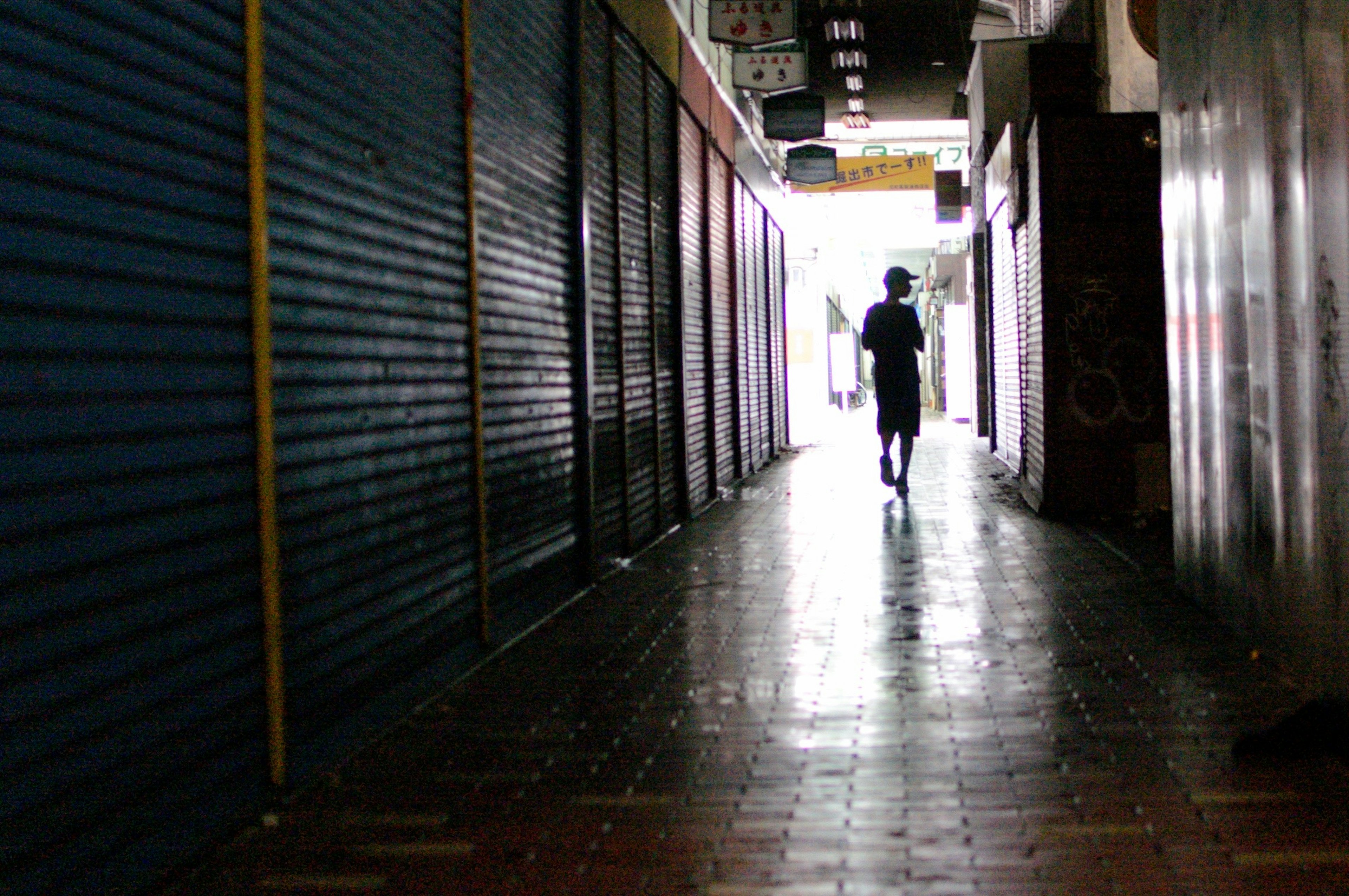 Silhouette of a person walking in a dimly lit corridor with closed shop shutters