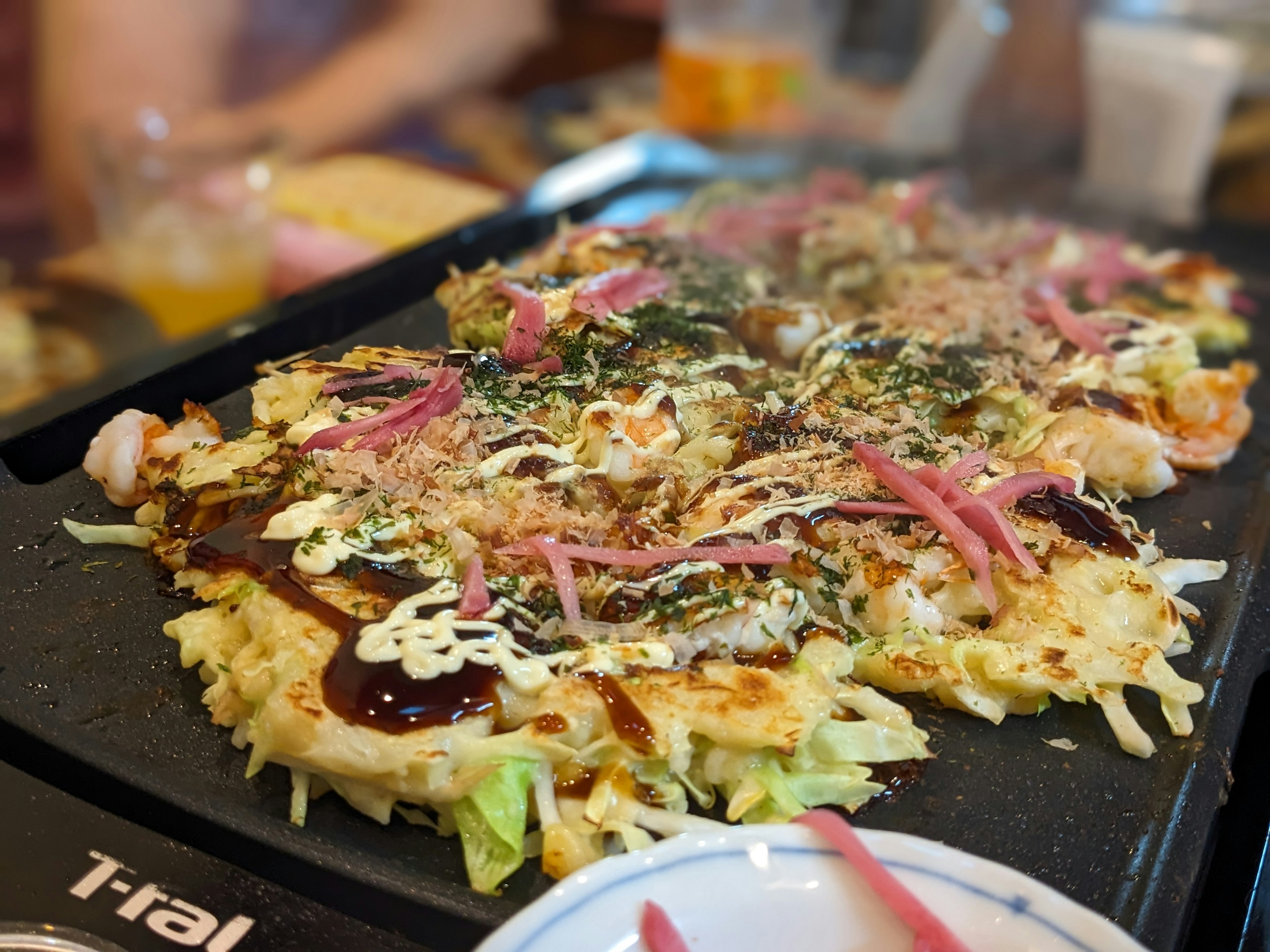 Okonomiyaki dimasak di atas piring panas, ditambahkan dengan jahe acar dan aonori