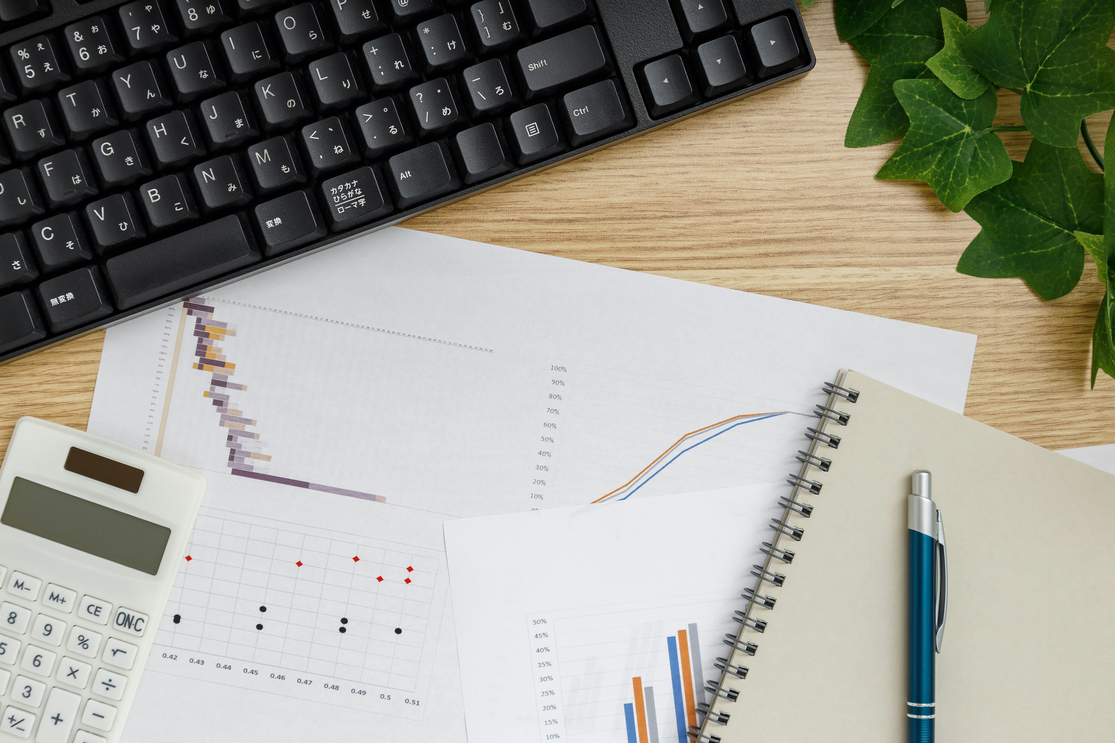 Un teclado, una calculadora y gráficos impresos sobre un escritorio con un cuaderno y un bolígrafo junto a una planta en maceta