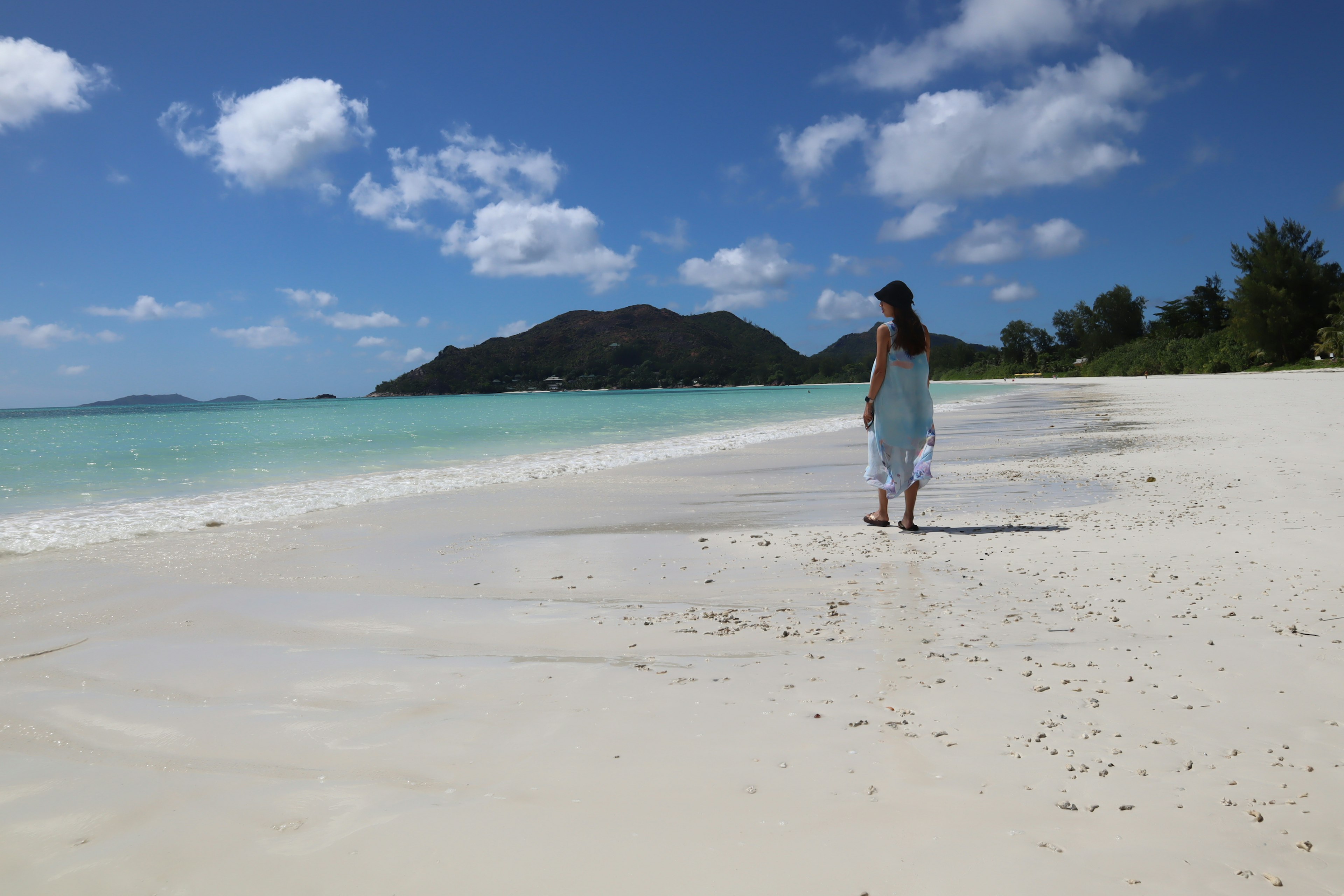 Woman walking along a white sandy beach with blue ocean and clouds