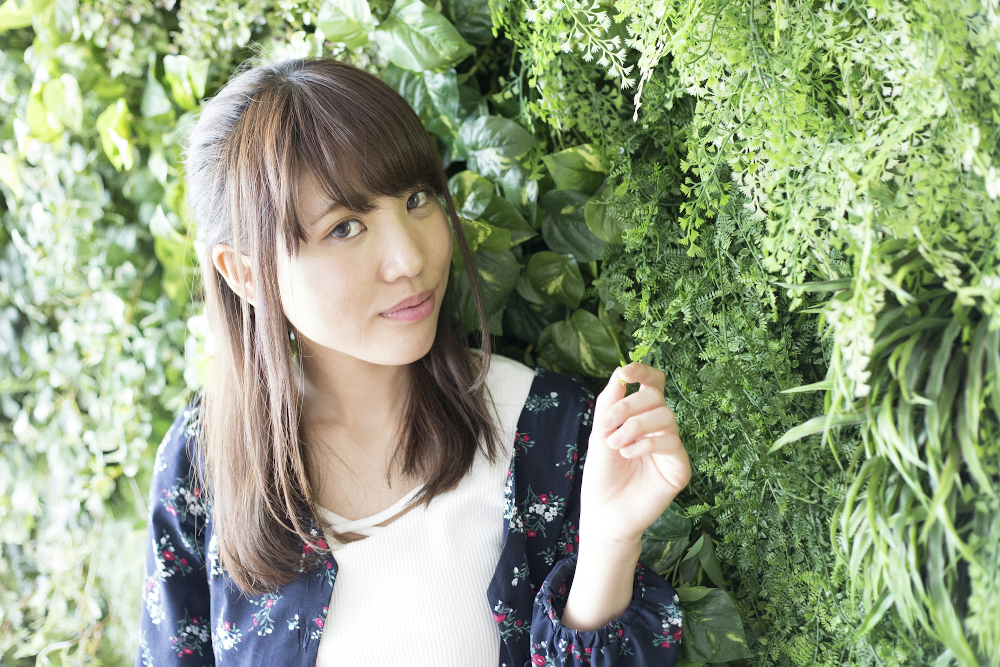 A woman standing in front of a green wall with various plants Long brown hair wearing floral clothing