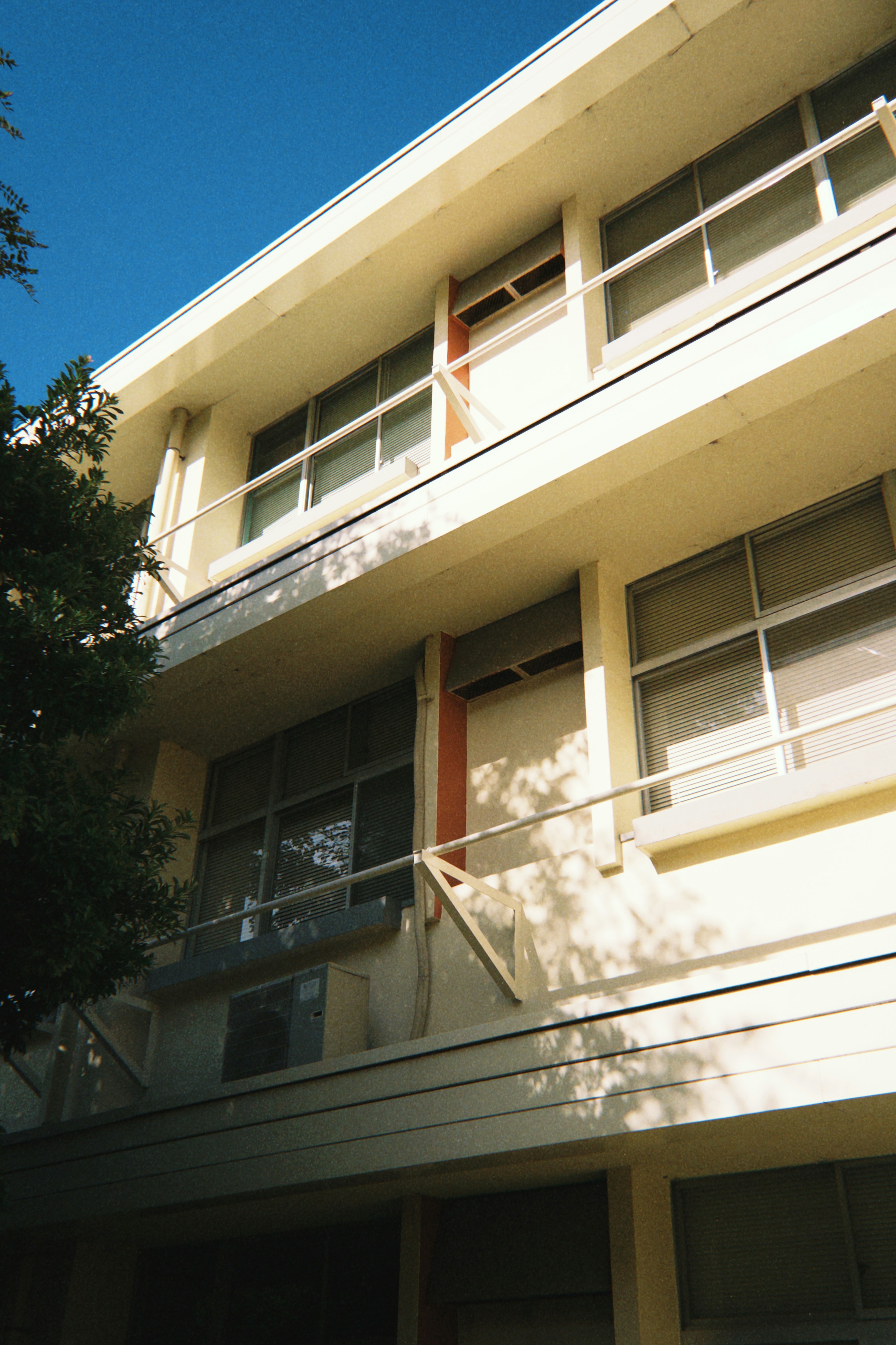 Extérieur d'un bâtiment de deux étages avec un ciel bleu lumineux, des reflets de fenêtres et des ombres d'arbres