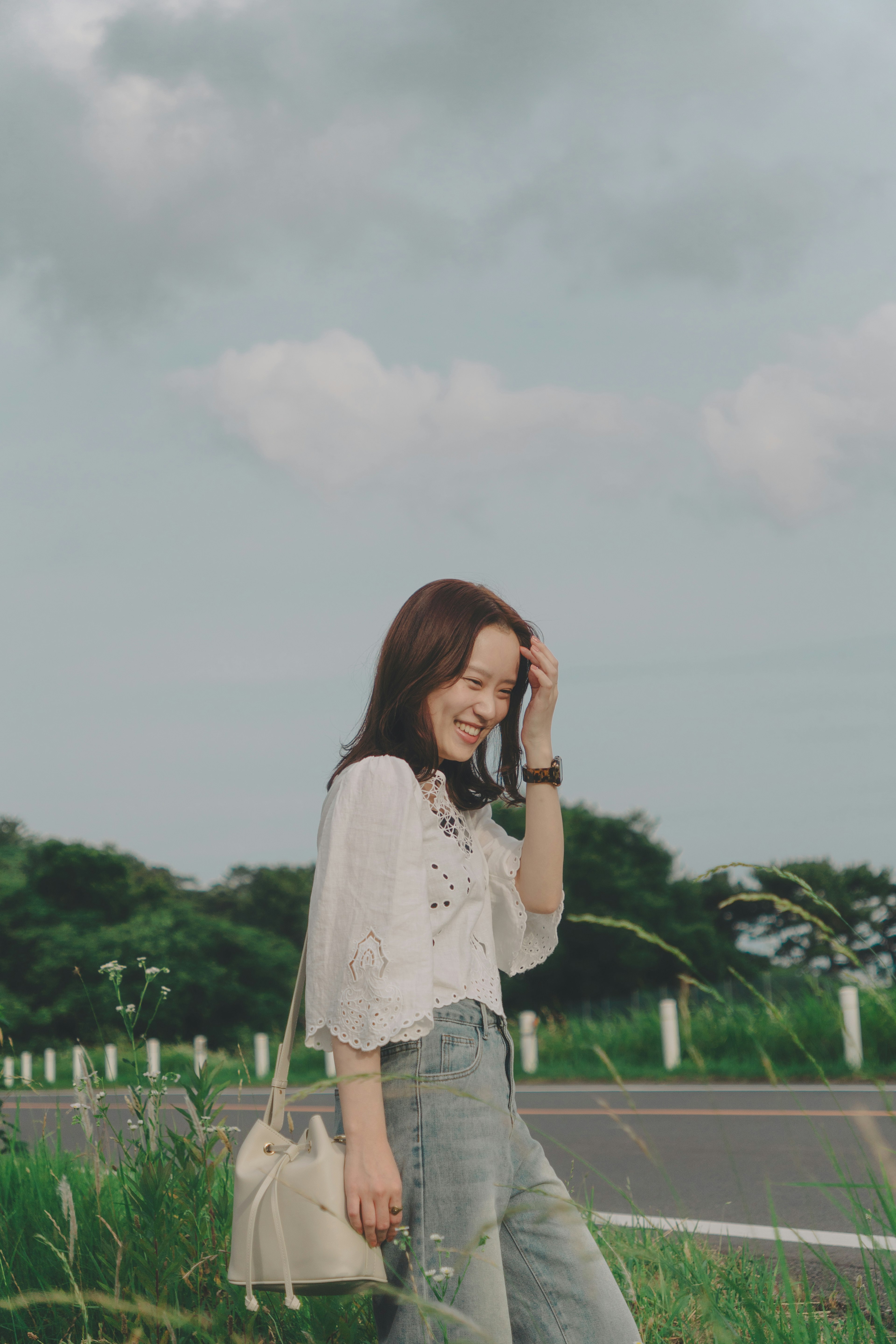 Mujer sonriente en un paisaje sereno vestida con una blusa blanca y jeans de mezclilla rodeada de hierba verde y una carretera