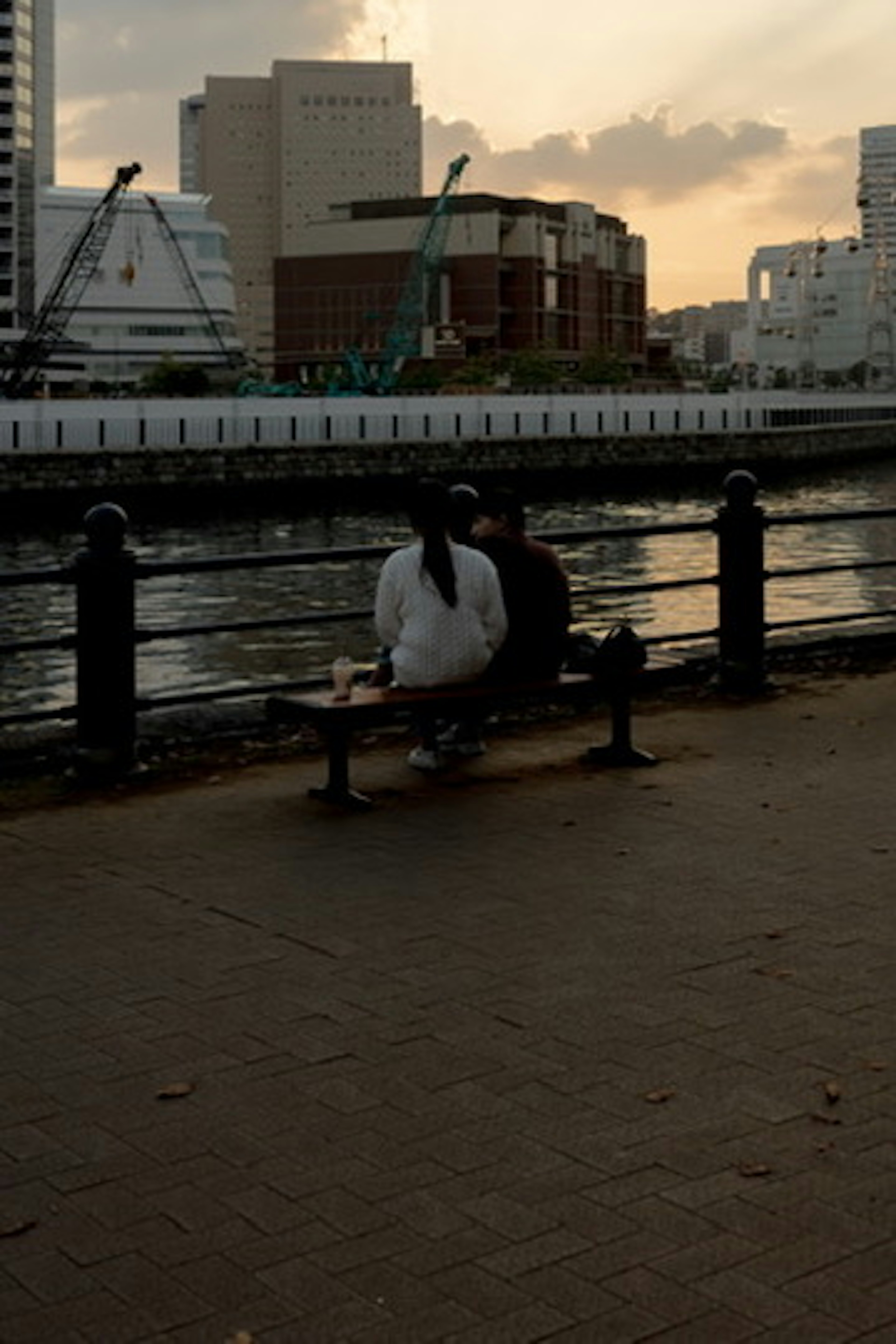 Two people sitting on a bench by the river at sunset with a city background