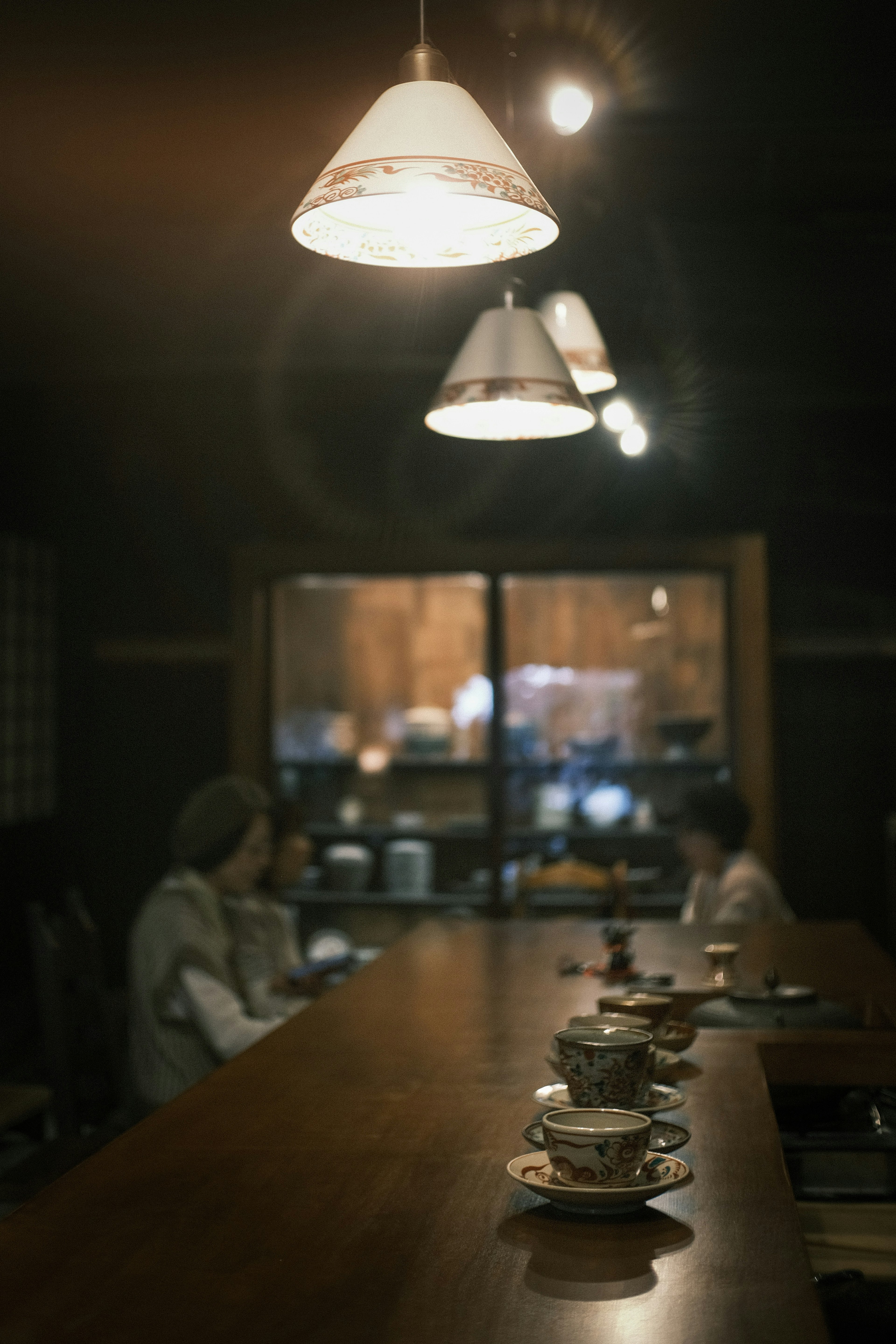 Interior of a dimly lit café with a long wooden counter featuring tea cups and pendant lights