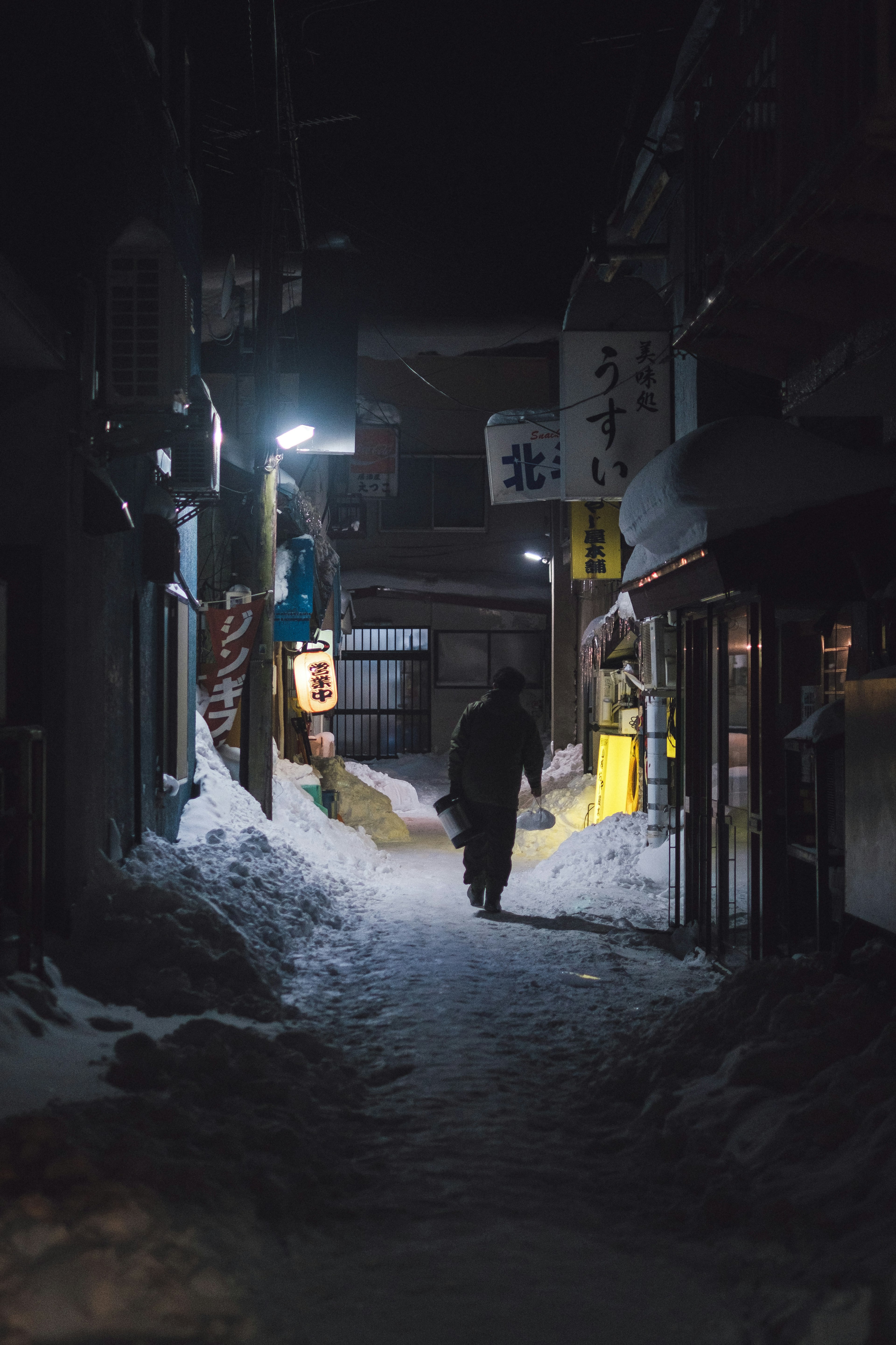 雪に覆われた夜の路地を歩く人物のシルエットと明かり