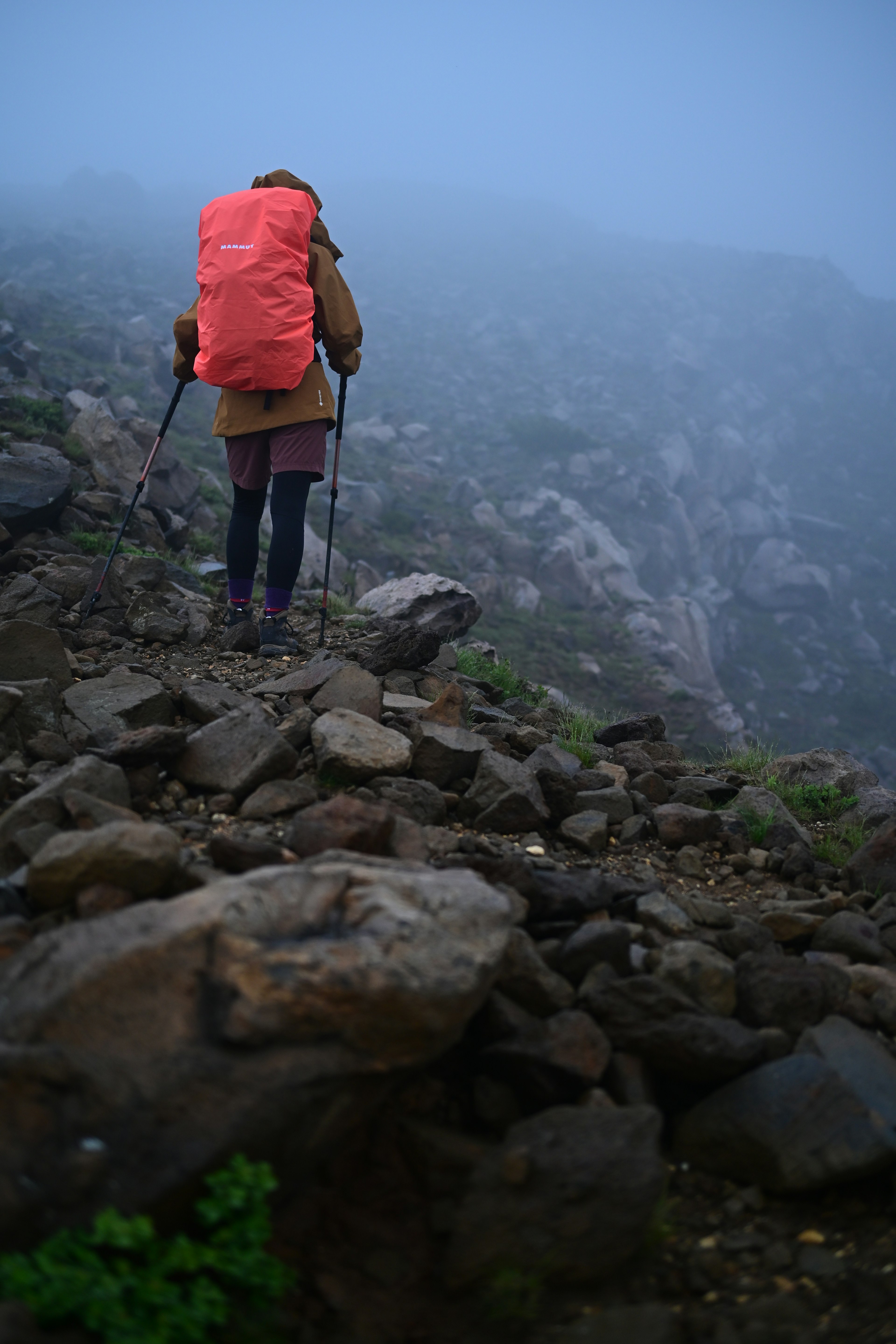 Hiker walking through fog carrying a large orange backpack navigating rocky terrain