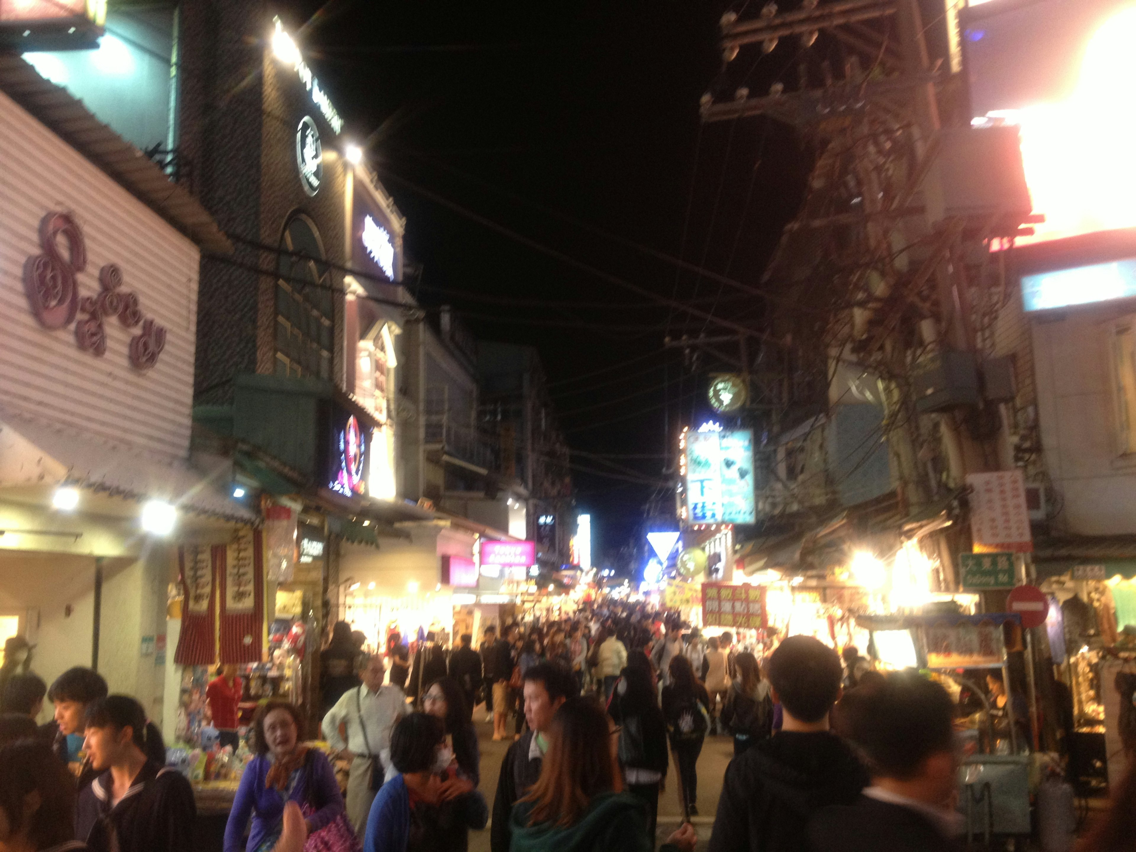 Calle de mercado nocturno concurrida llena de gente y letreros brillantes