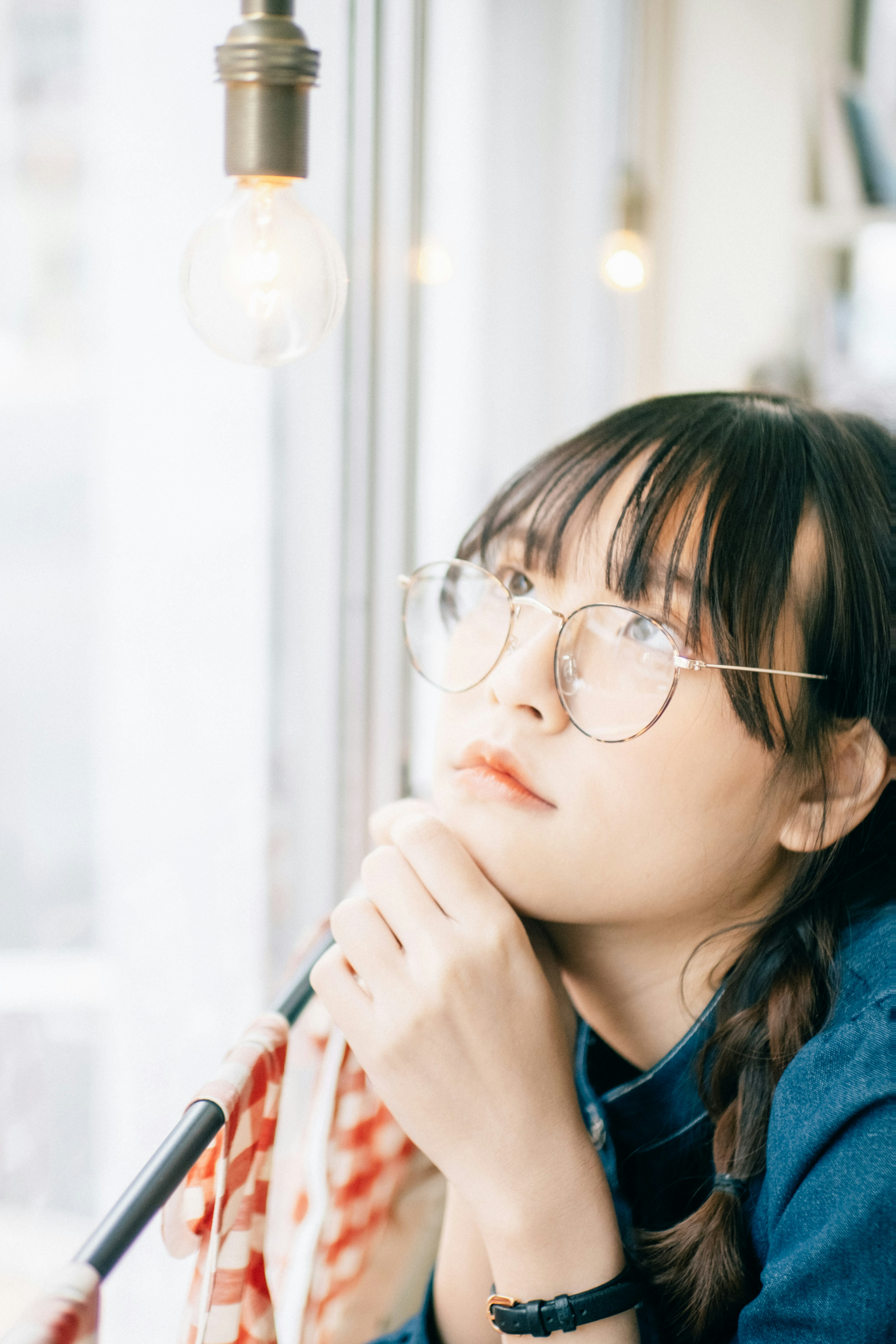 A young woman deep in thought by the window wearing glasses and braids with a warm light bulb above her