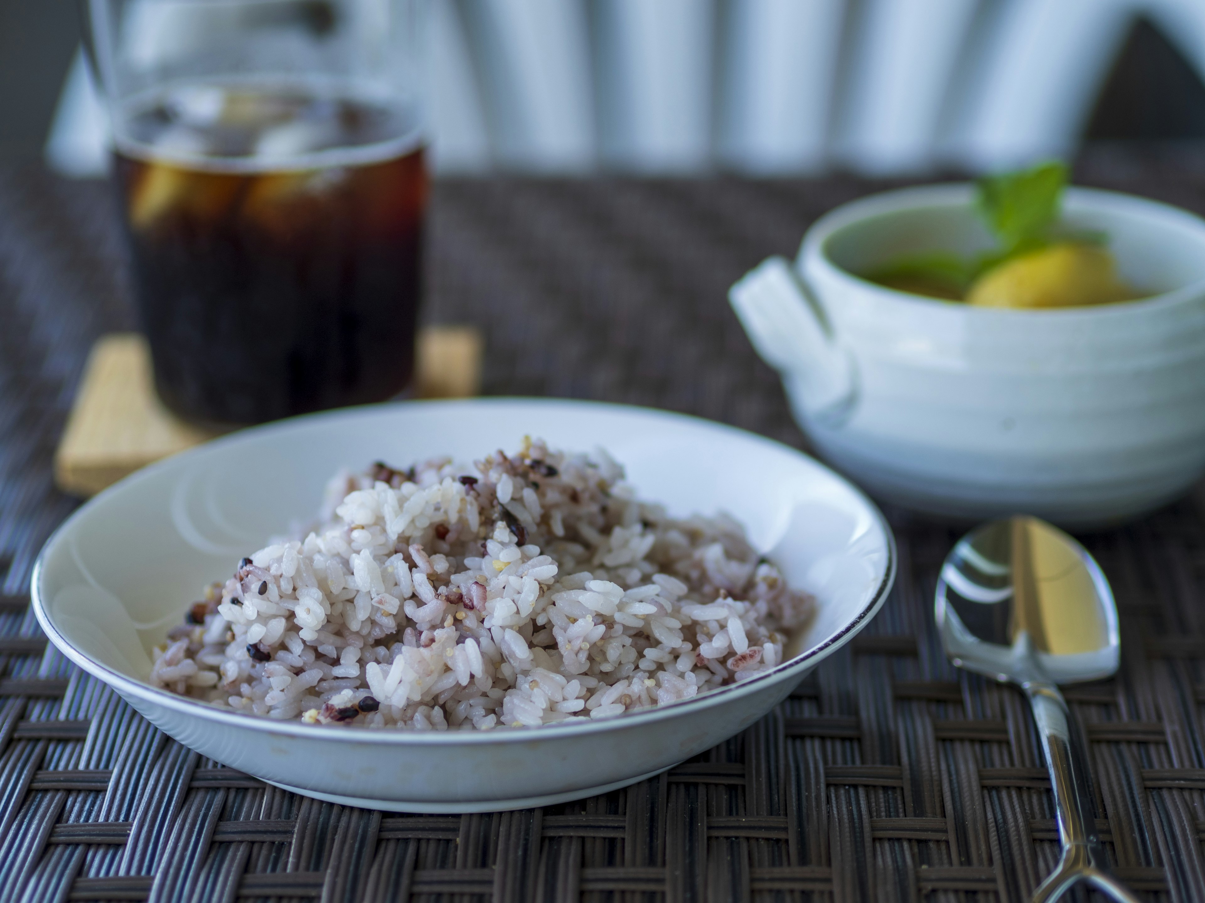 Bowl of mixed black and white rice with a glass of cold beverage and a small dish of fruit in the background