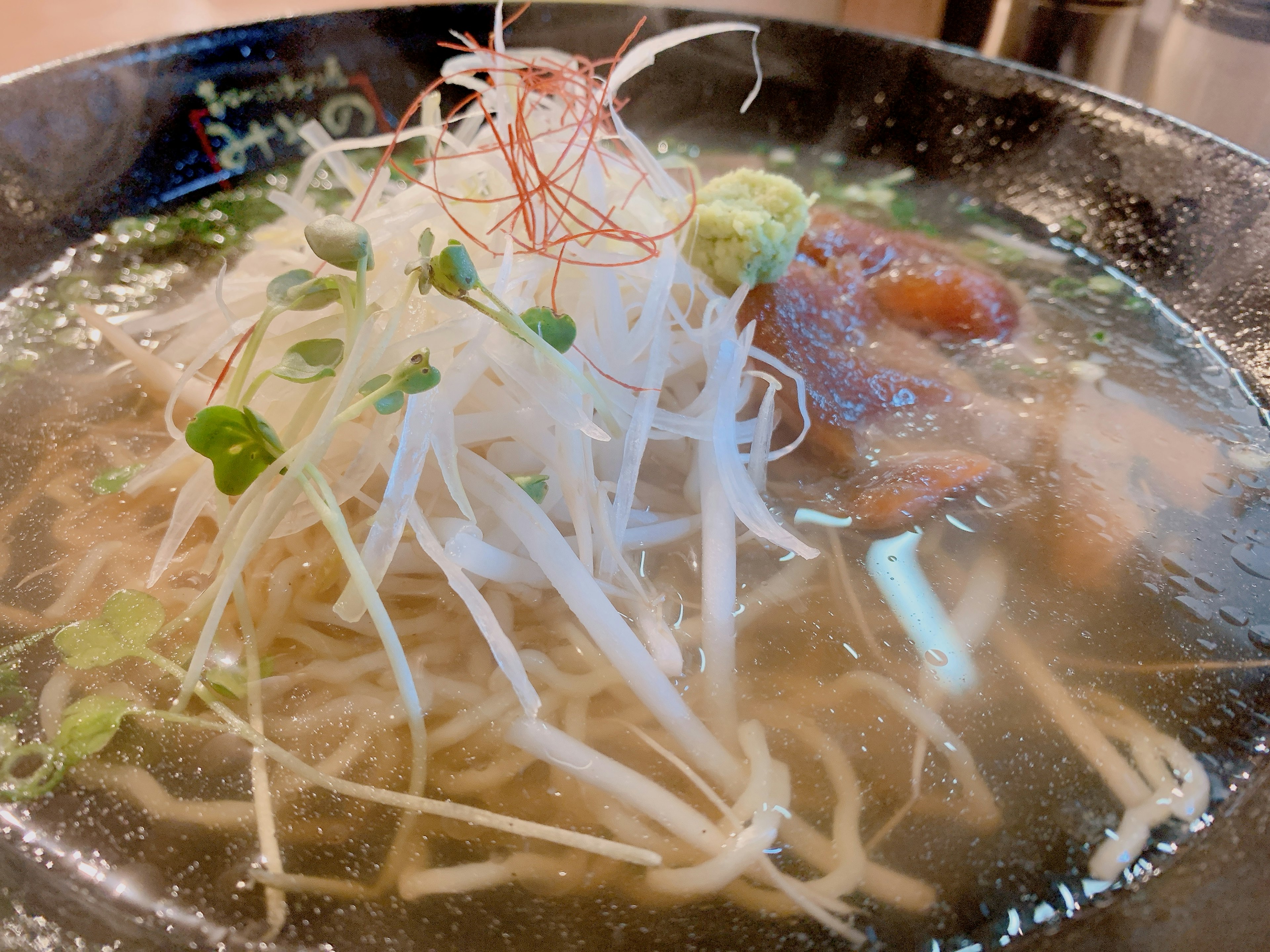 A bowl of clear broth ramen with thin noodles topped with white scallions and green herbs