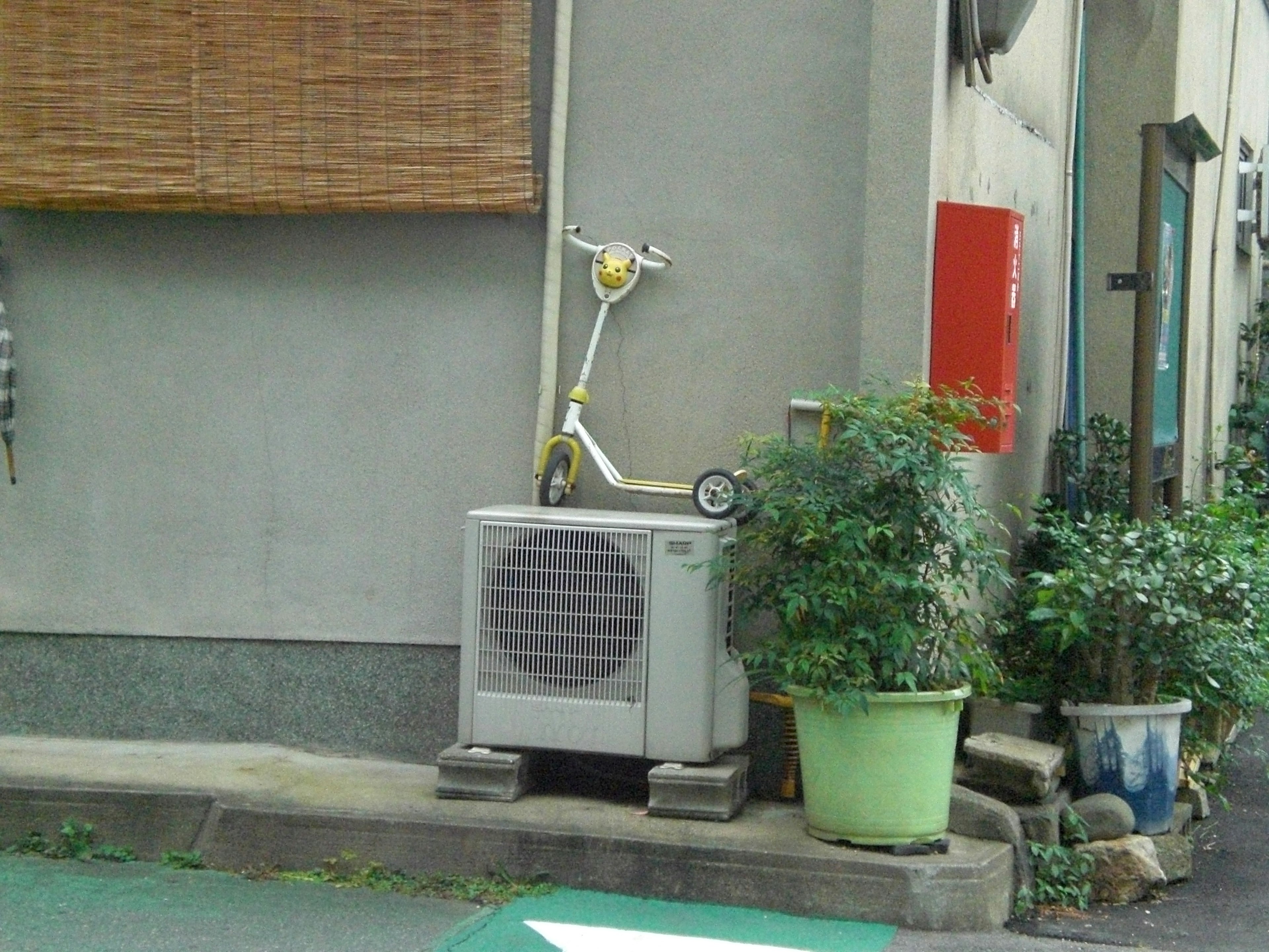 建物の側面に置かれたエアコンの上に乗った黄色いスクーターと緑の鉢植え