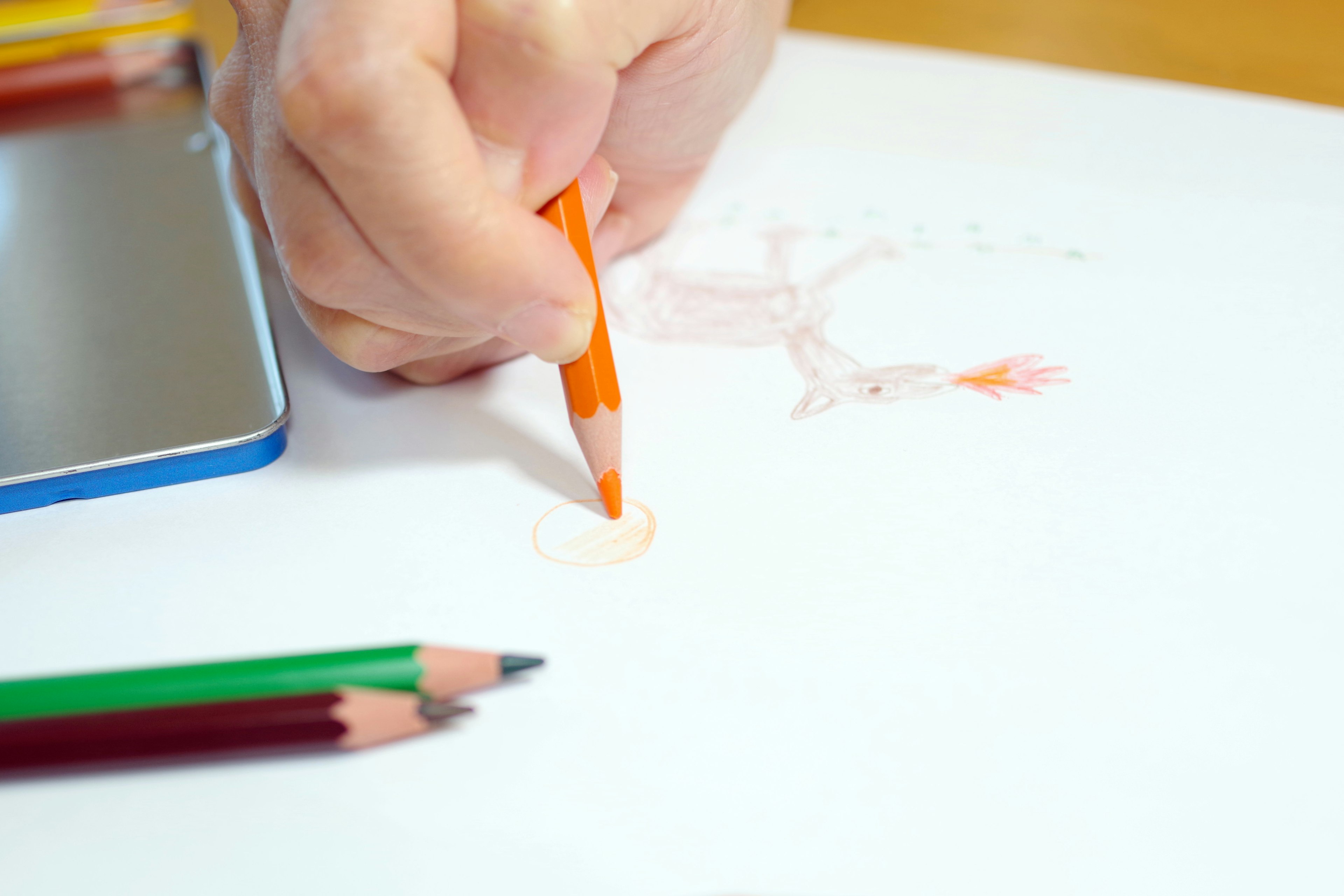 A hand using an orange colored pencil on white paper with green colored pencils nearby