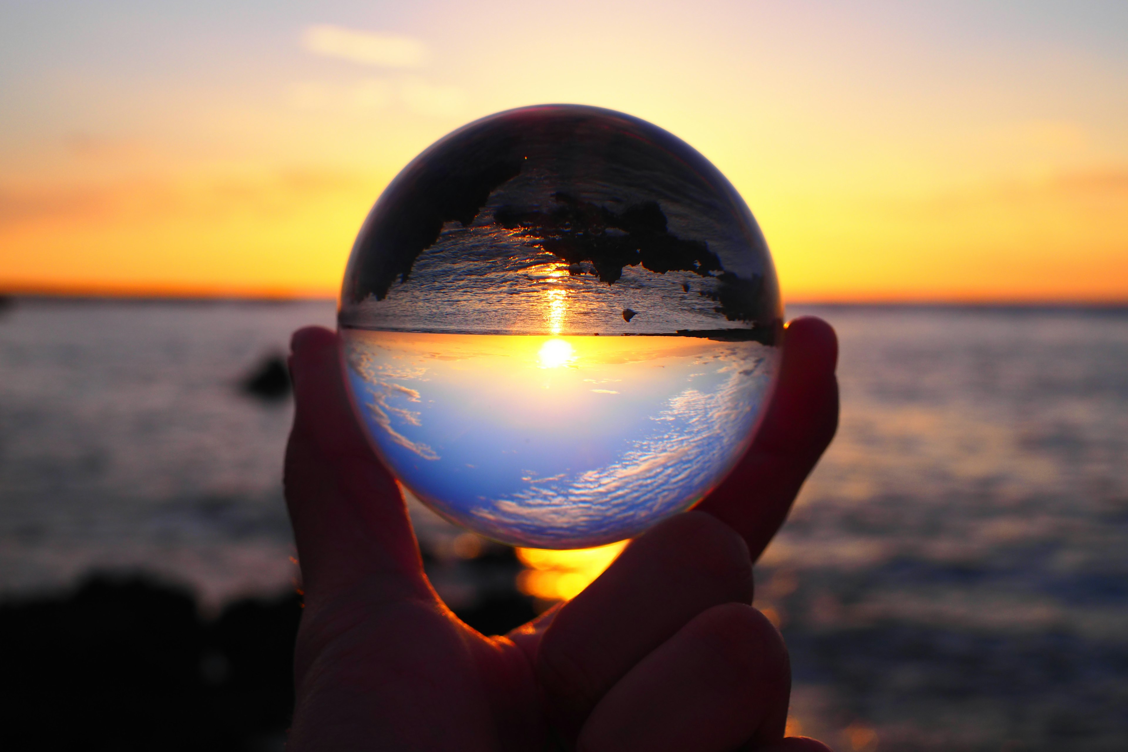 Una sfera di cristallo tenuta in mano che riflette il tramonto e la vista sull'oceano