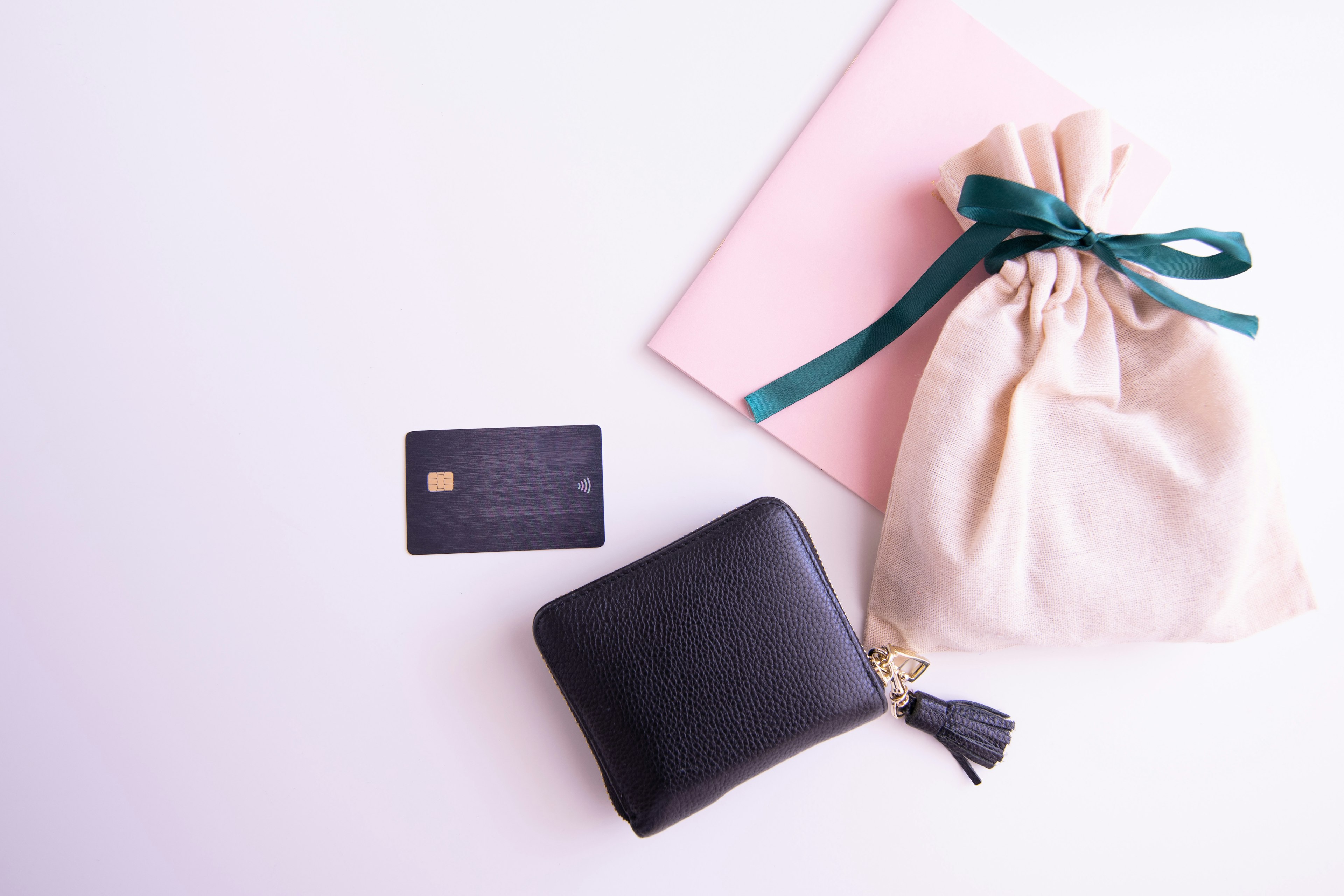 Un portefeuille noir et une carte de crédit à côté d'un carnet rose et d'un sac en tissu blanc noué avec un ruban vert sur une surface plate