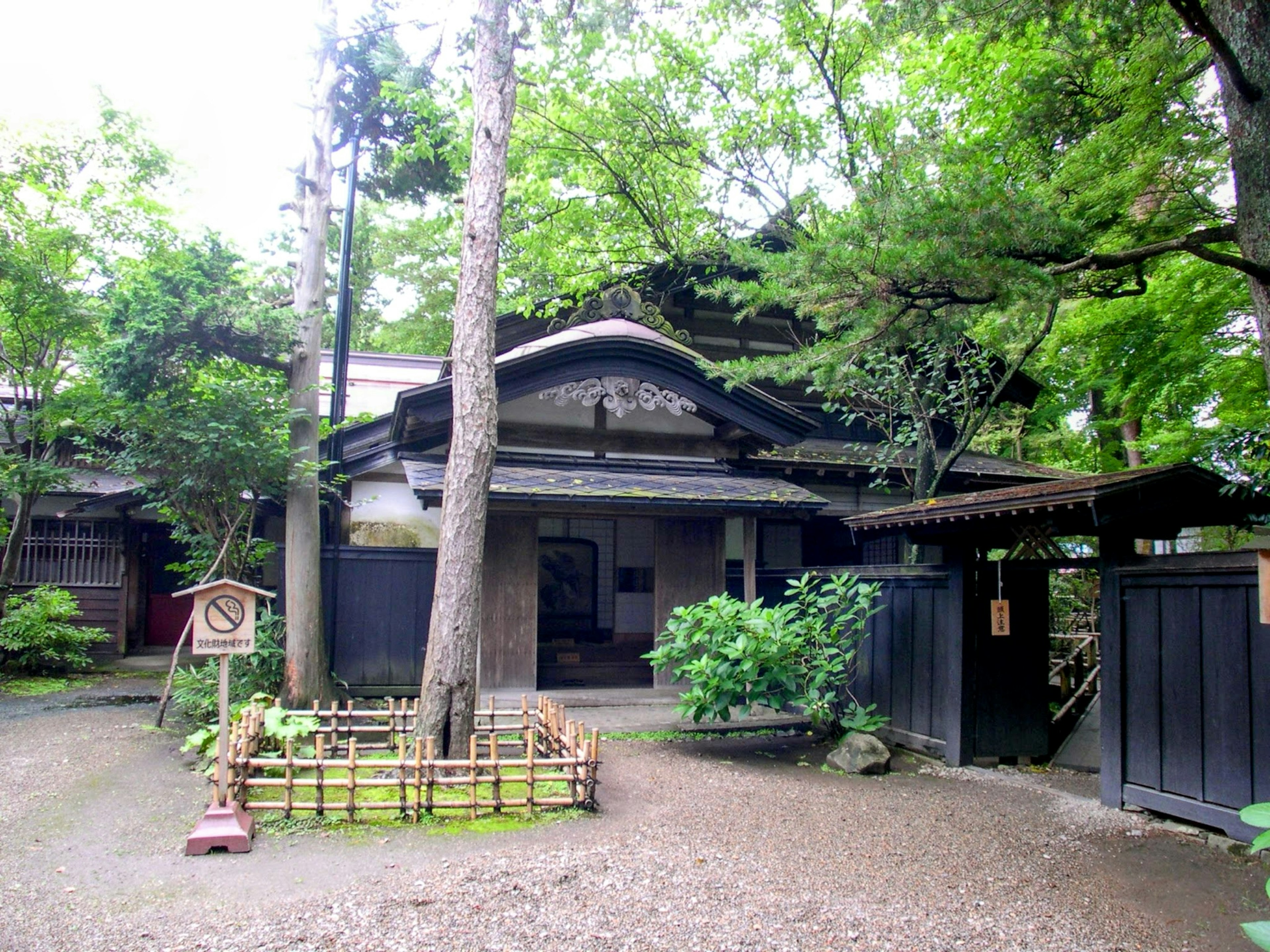 Exterior de una casa tradicional japonesa rodeada de vegetación, cerca de madera, diseño de techo distintivo