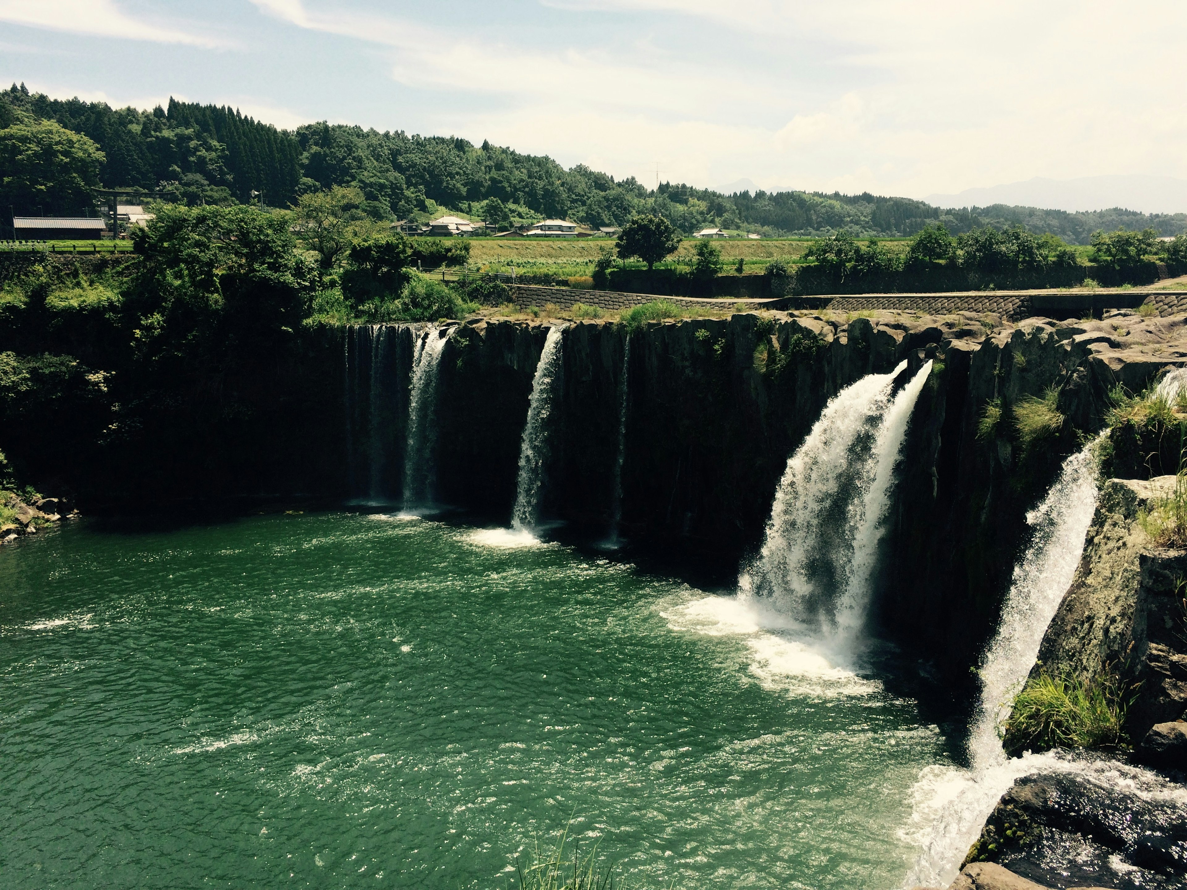 Vue panoramique de chutes d'eau tombant dans une eau verte entourée de verdure luxuriante et d'un ciel bleu