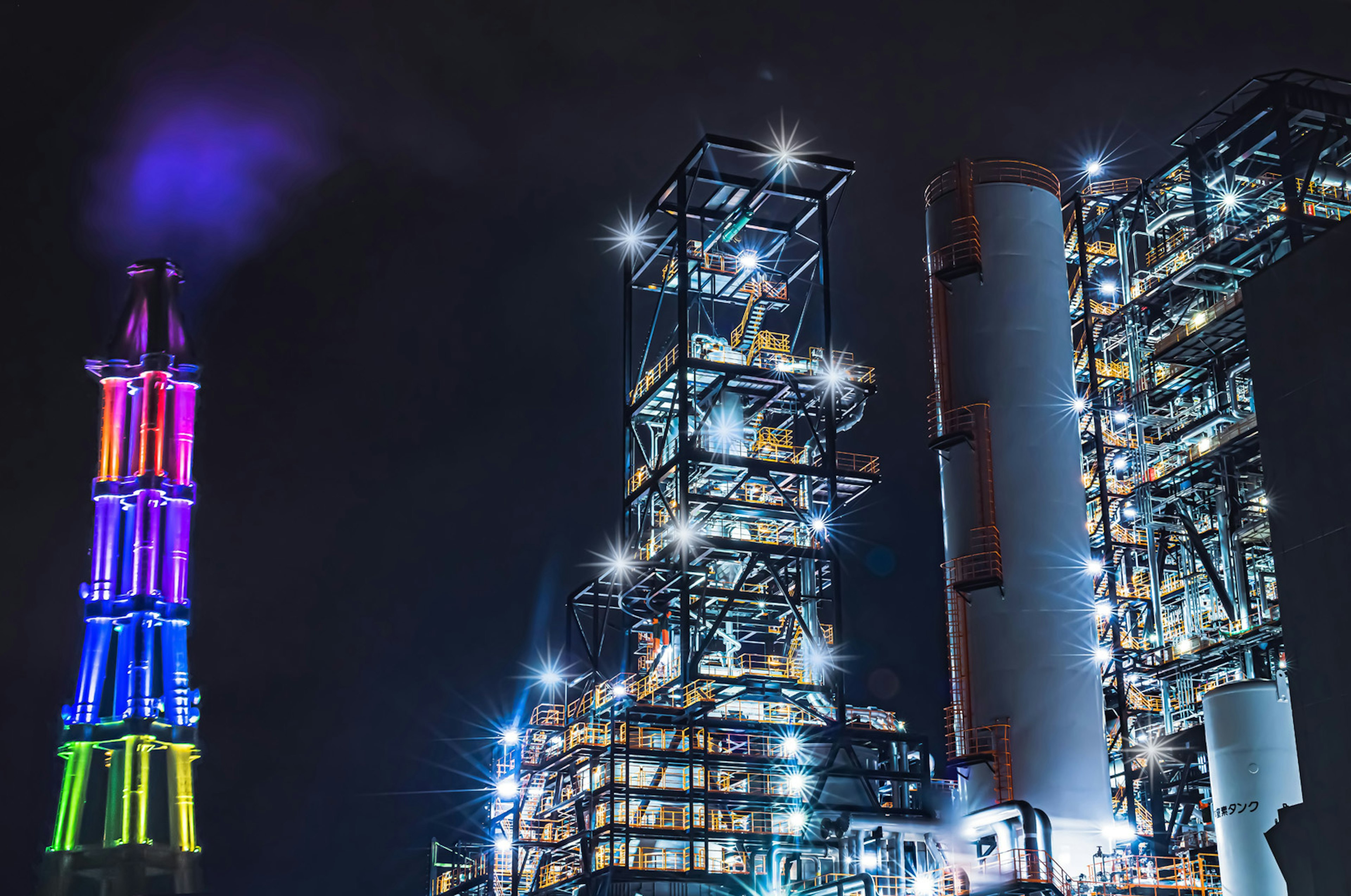 Illuminated industrial plant at night featuring colorful smokestack and structures