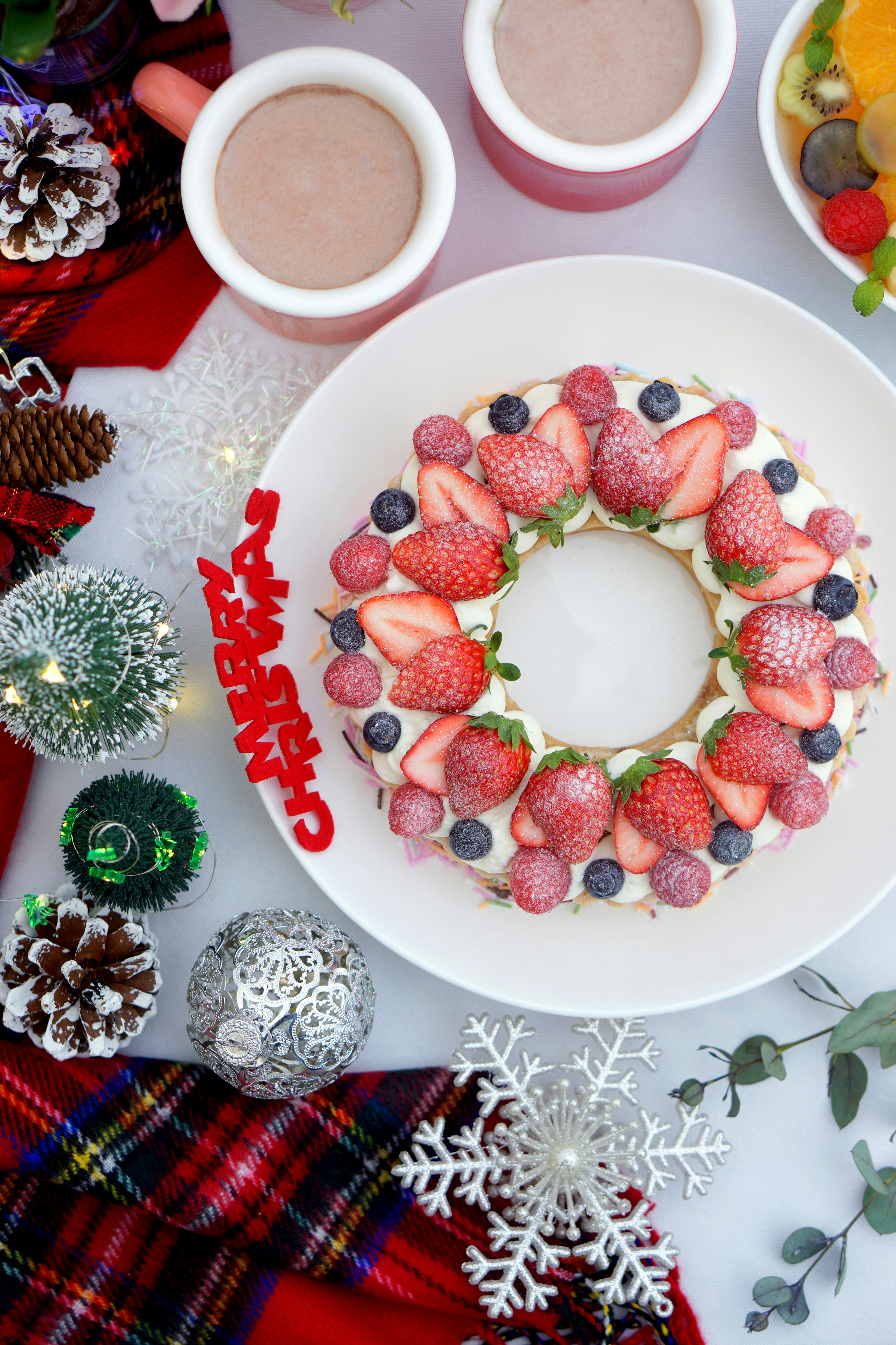 Plato de postre navideño decorado con fresas y arándanos en forma de corona con tazas de chocolate caliente y frutas variadas