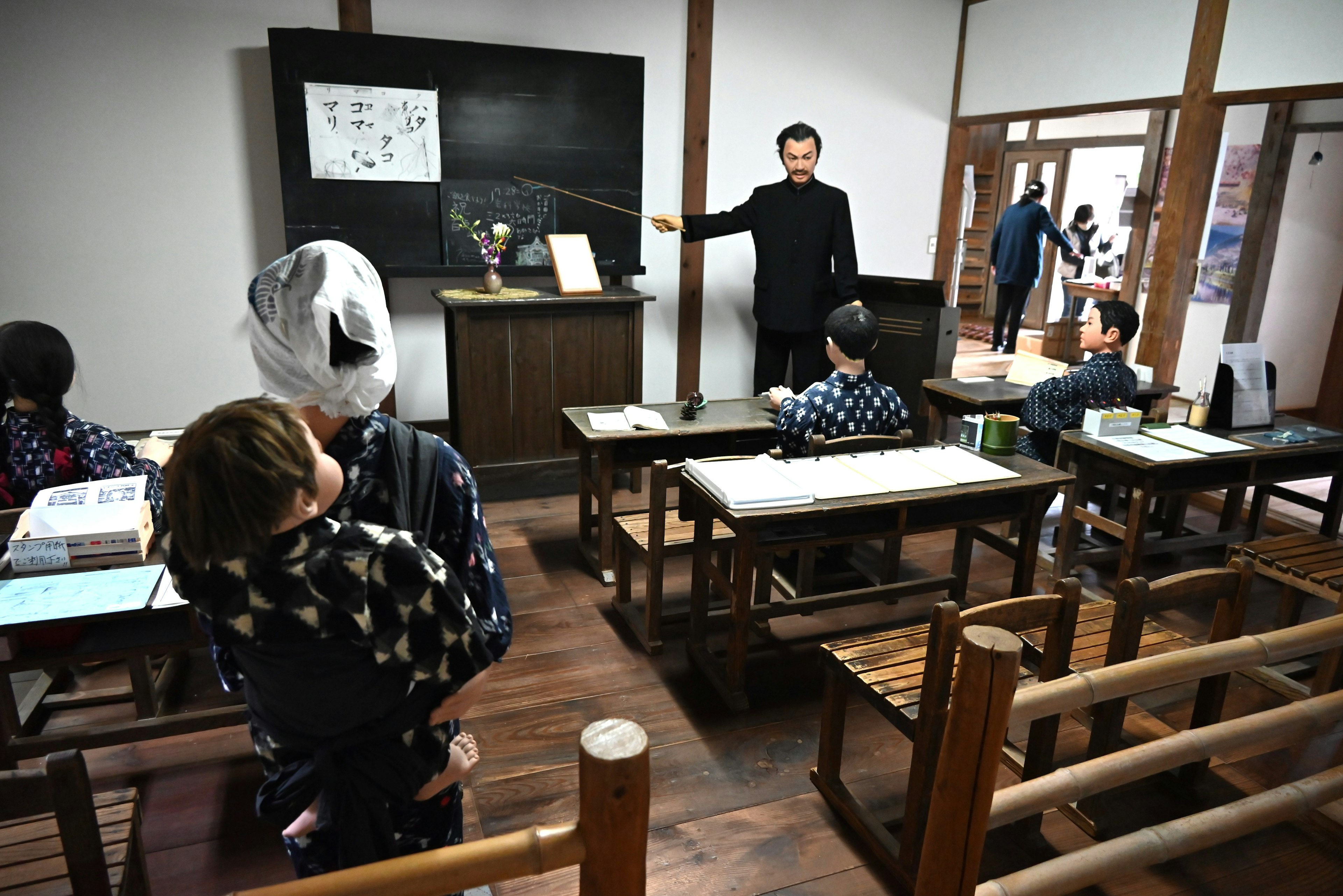 Escena de aula con un maestro señalando una pizarra estudiantes sentados en escritorios