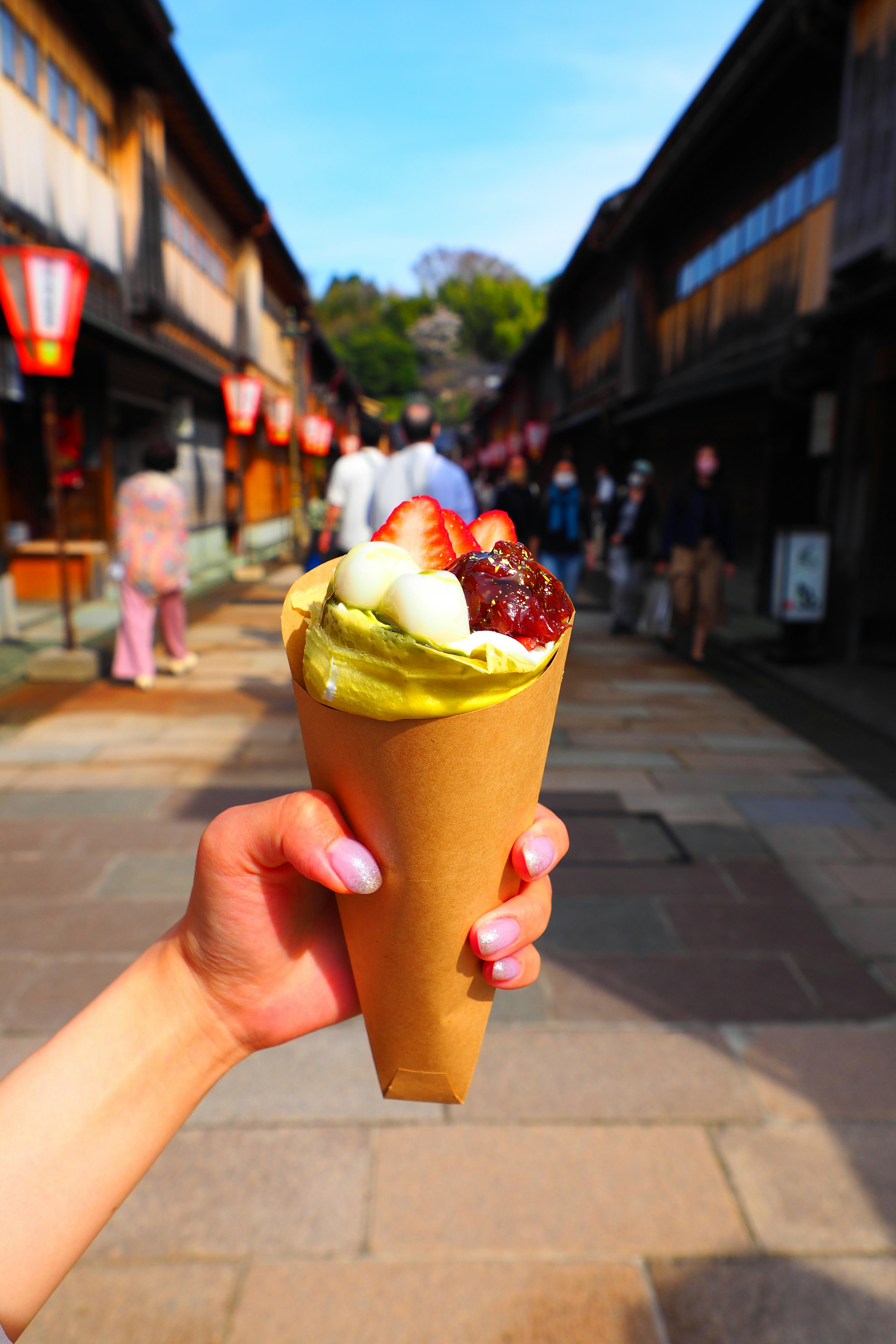 A hand holding a matcha ice cream and fruit dessert with a traditional street in the background