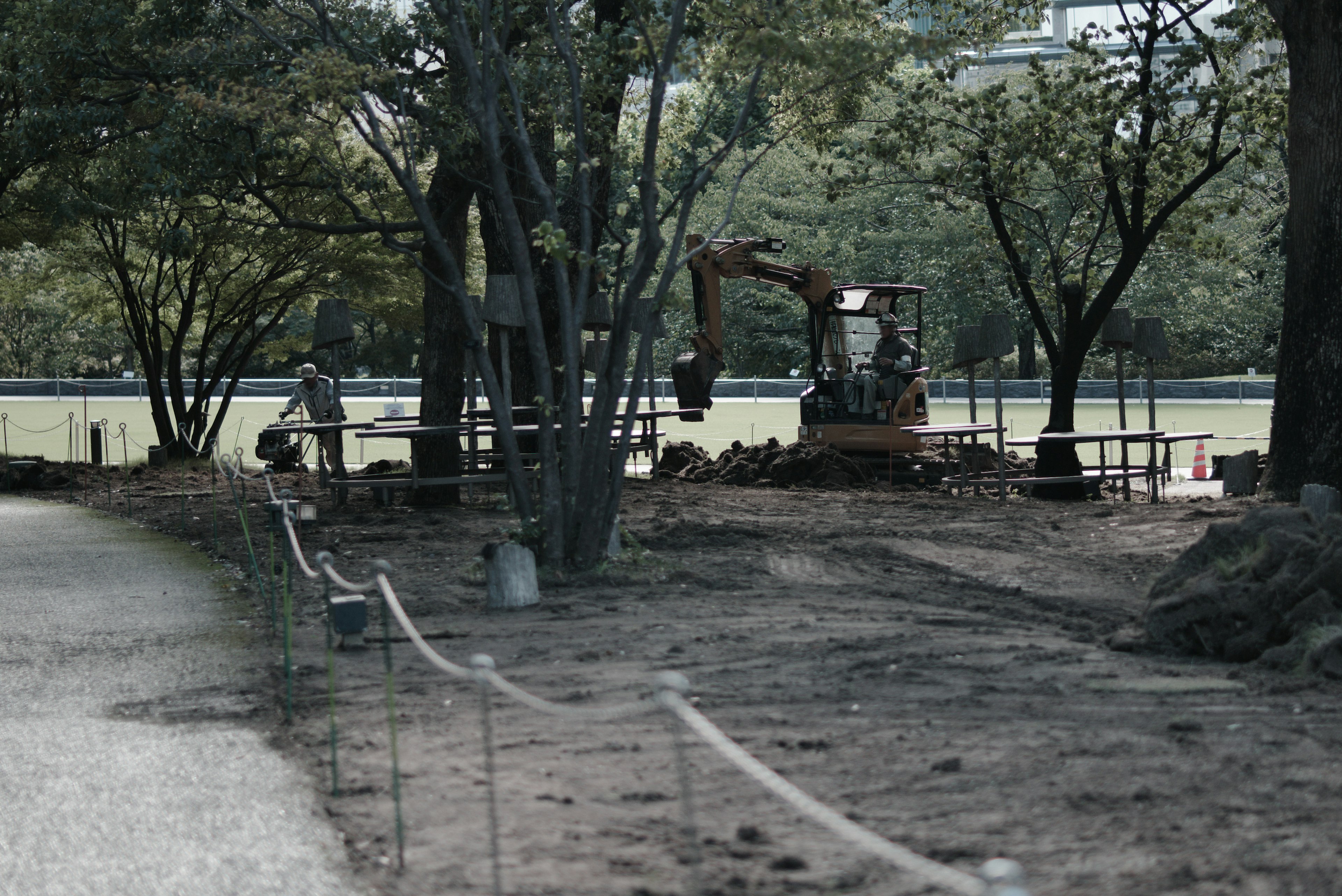 Construction site in a park with machinery and workers visible green trees and disturbed earth