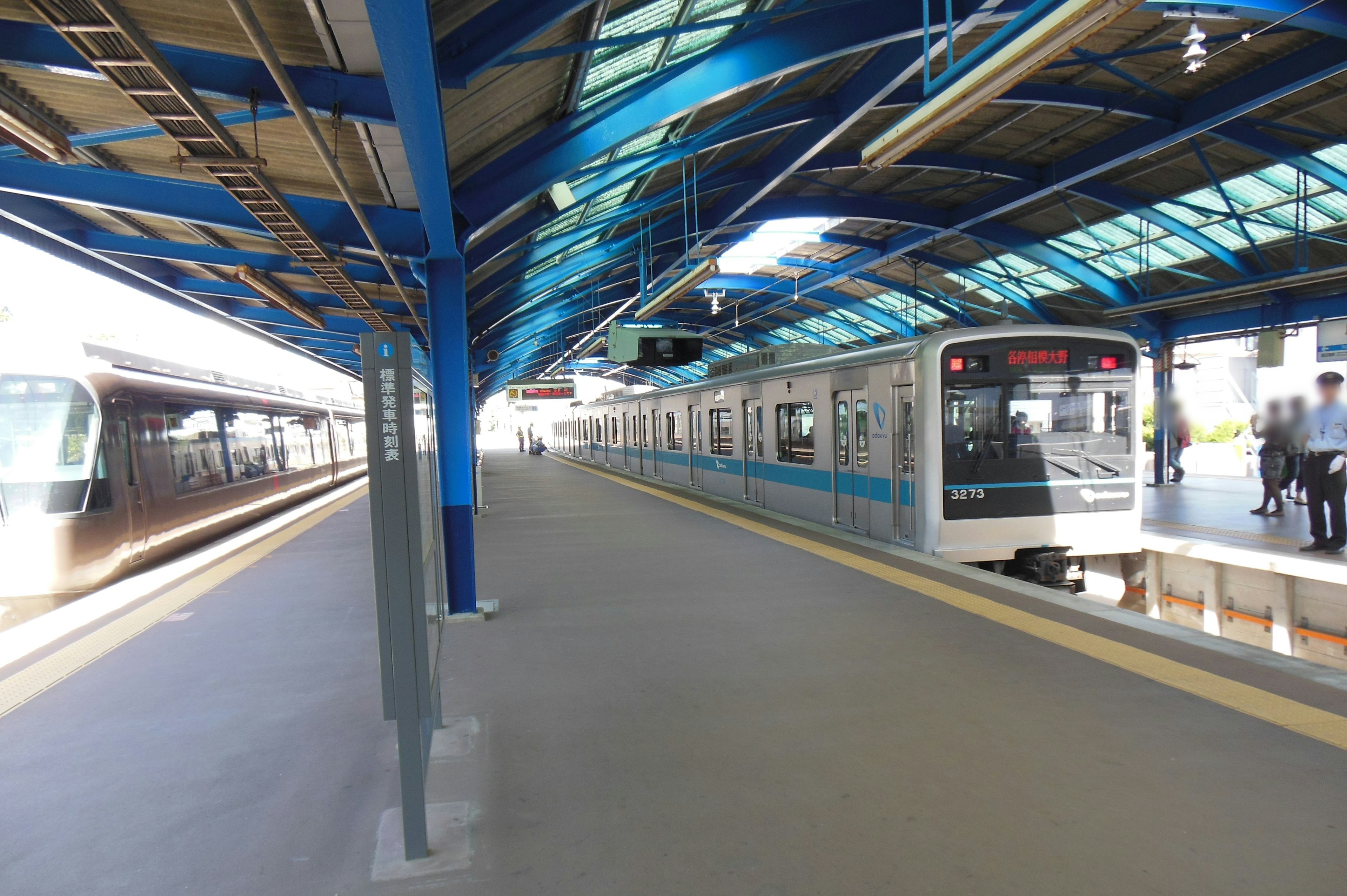 Train at a blue-roofed station platform with people and bright sunlight