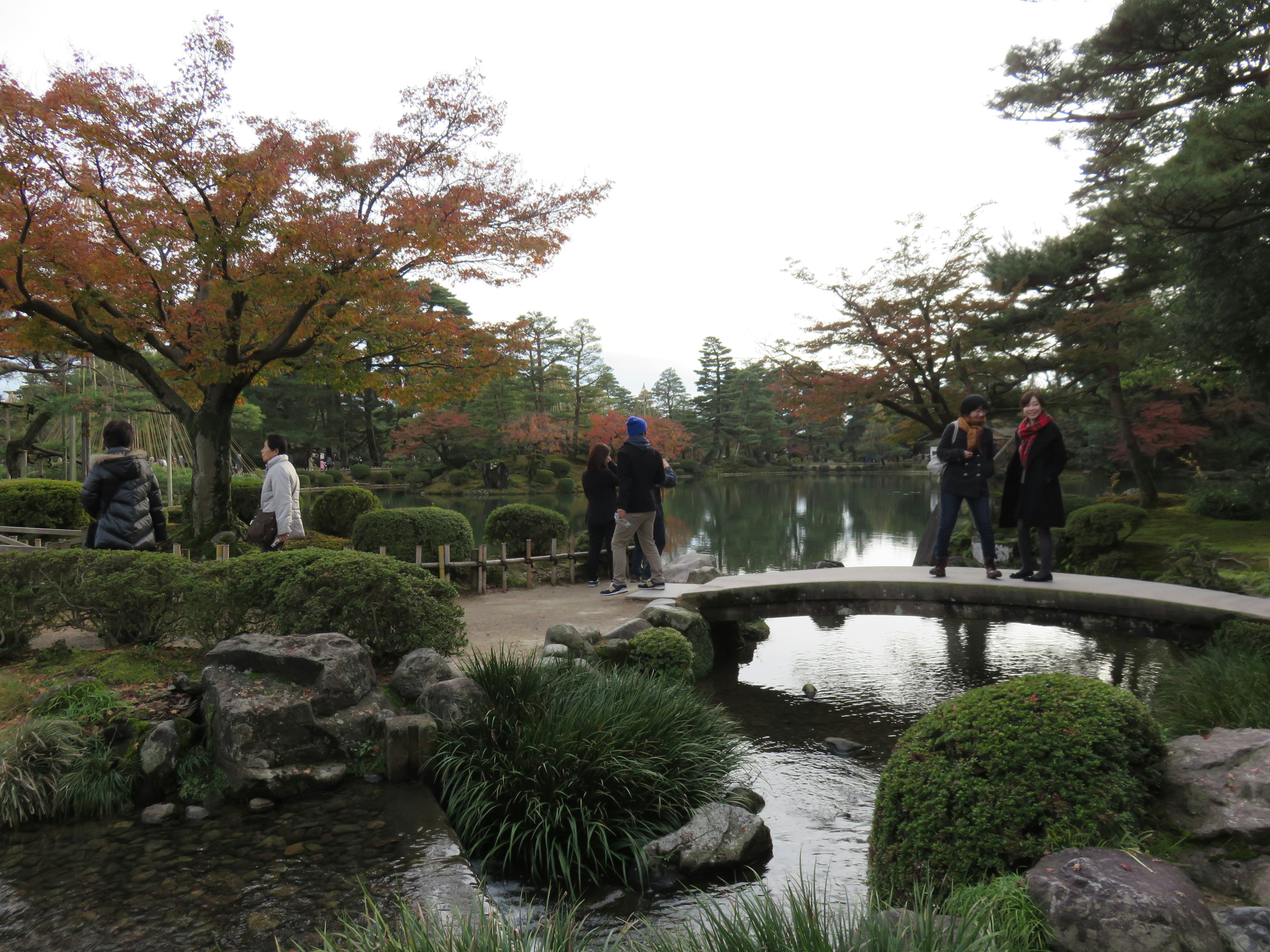 Malersicher japanischer Garten im Herbst mit buntem Laub Menschen, die eine Steinbrücke überqueren und einem ruhigen Teich