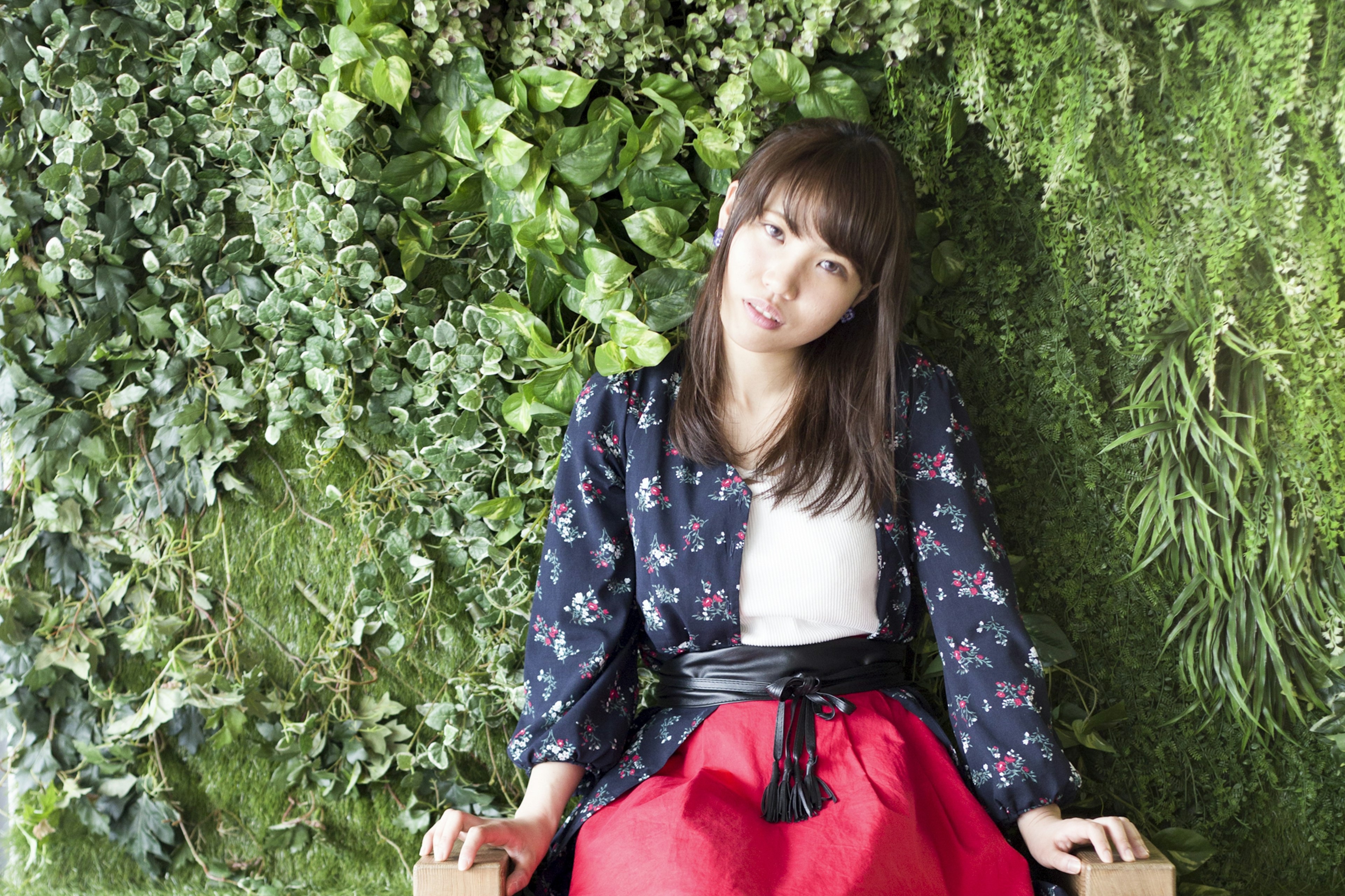 A woman sitting in front of a green wall wearing a floral jacket and a red skirt