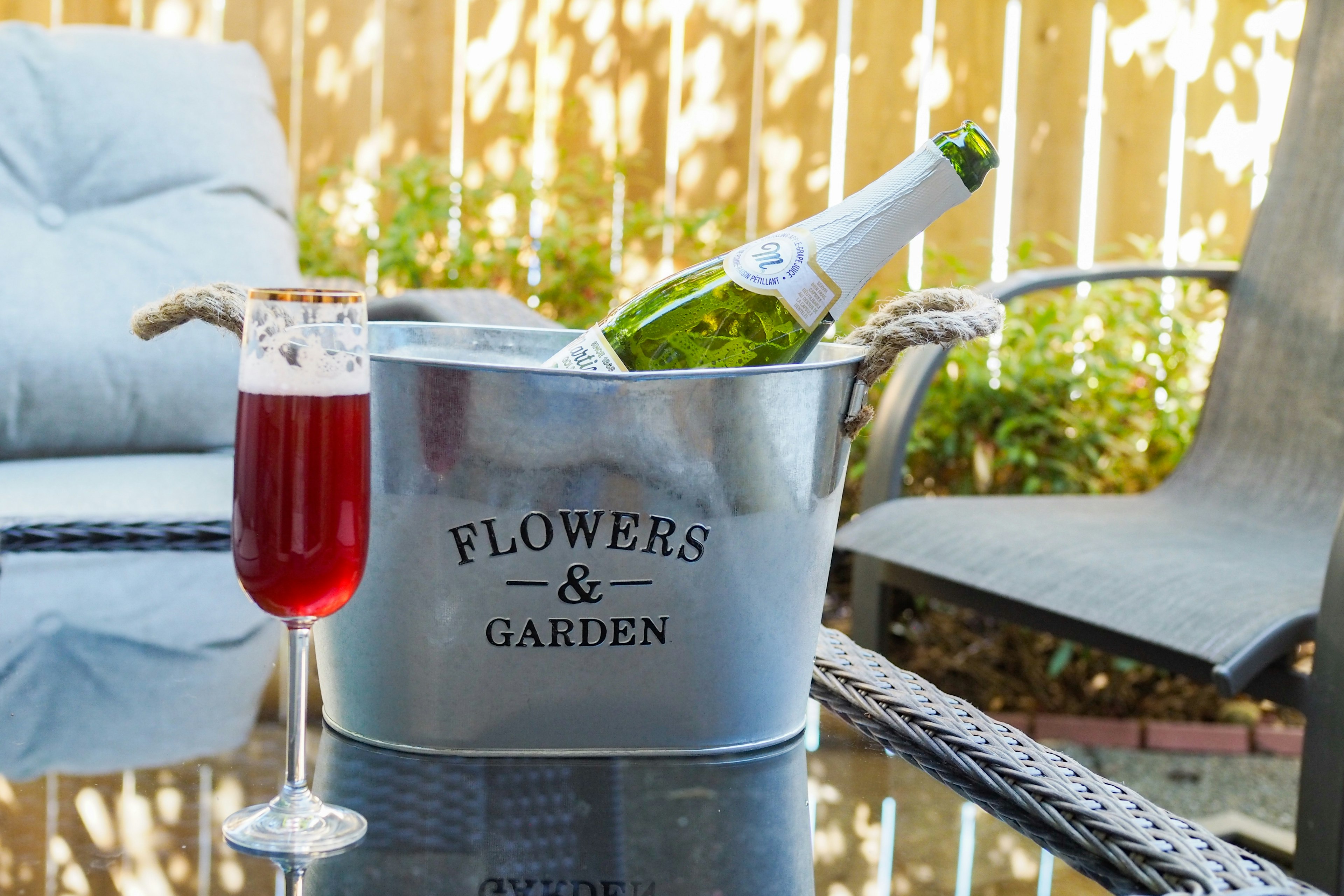 Outdoor scene featuring a champagne bottle in a floral garden bucket and a red cocktail glass