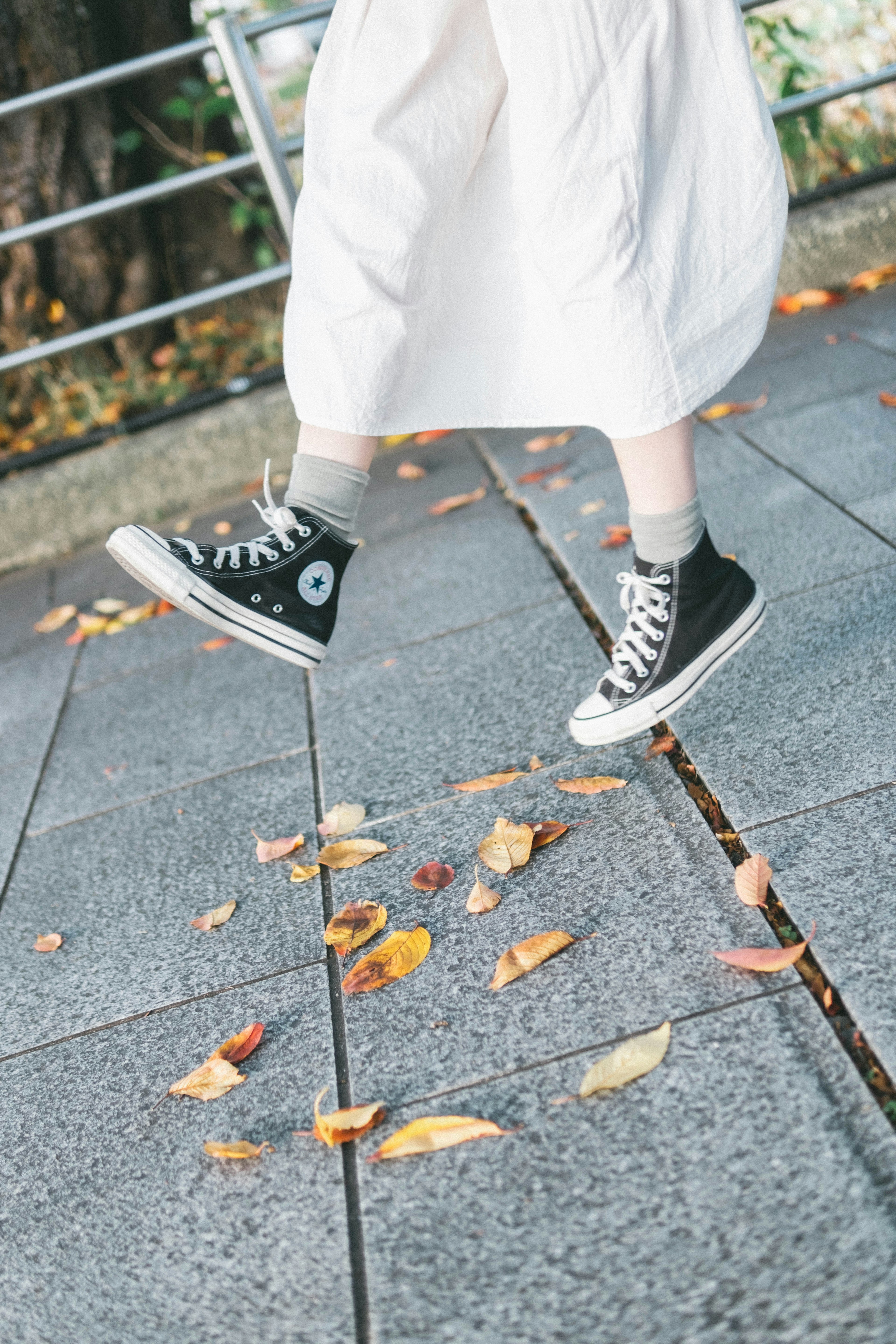 Person in a white skirt walking in black sneakers with fallen leaves on the ground