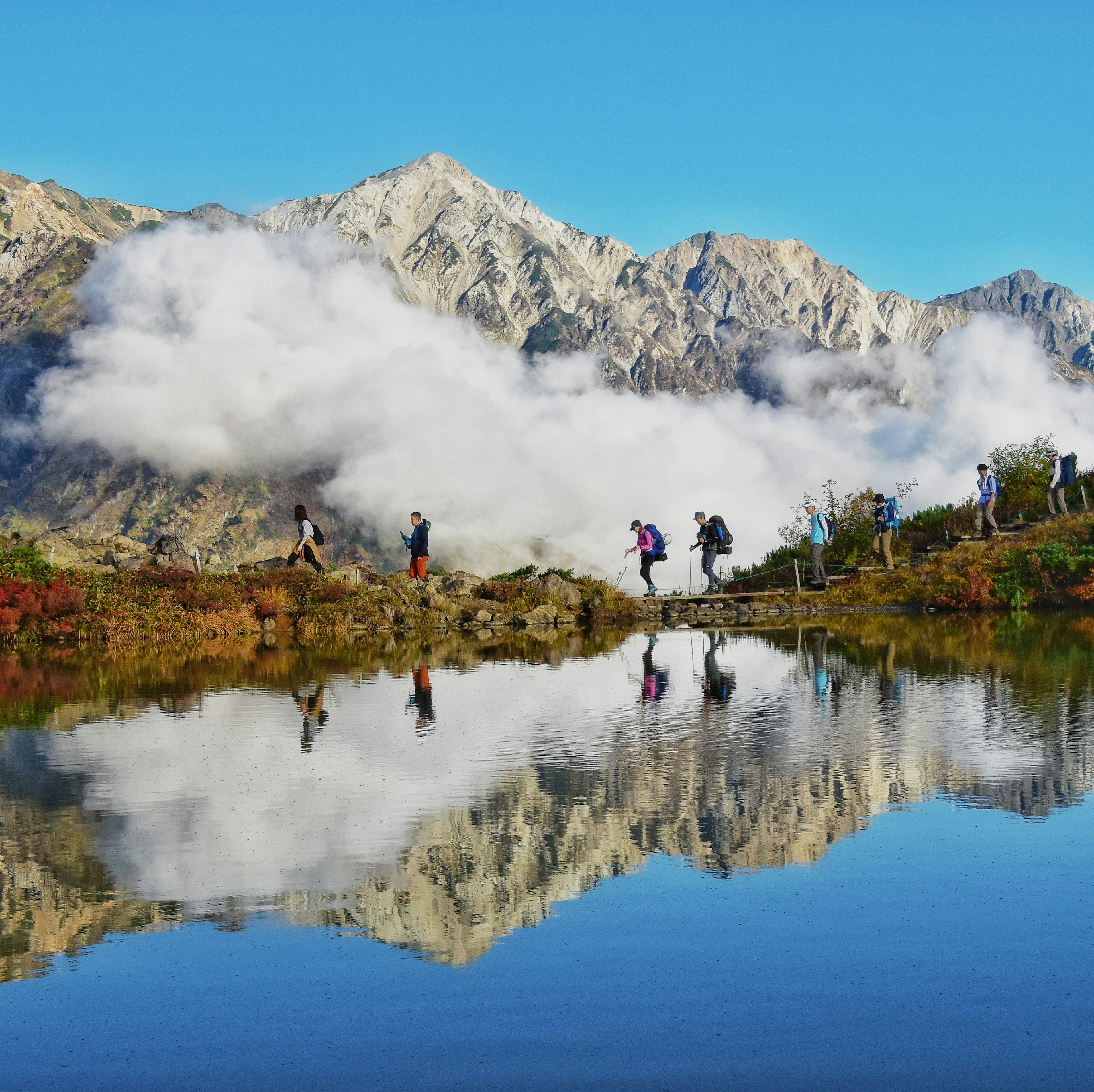 Scenic view of hikers walking under beautiful mountains and blue sky with clouds reflecting on the water surface