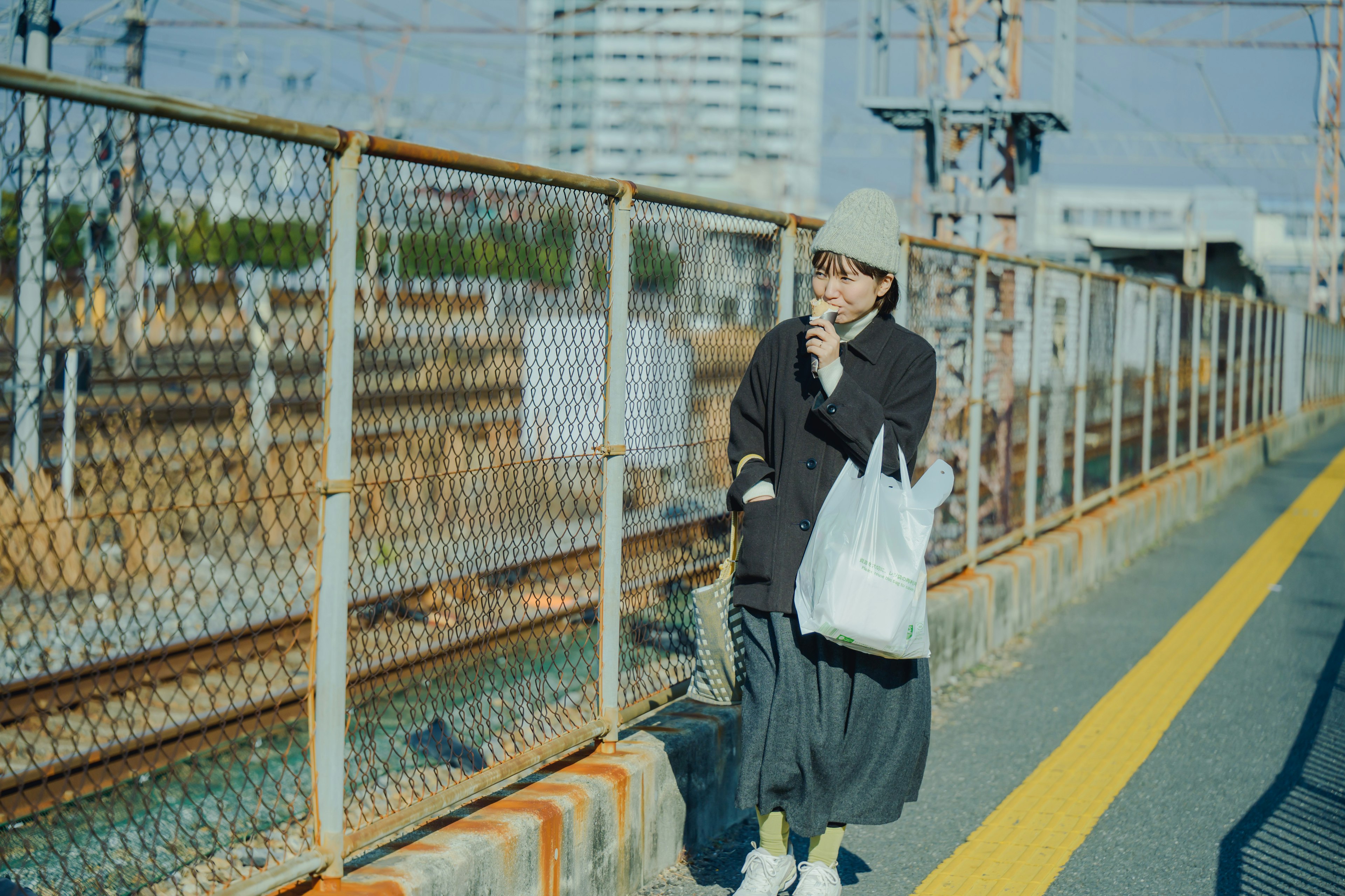 Young woman standing on a train platform smoking a cigarette wearing a black coat and gray skirt holding a white bag