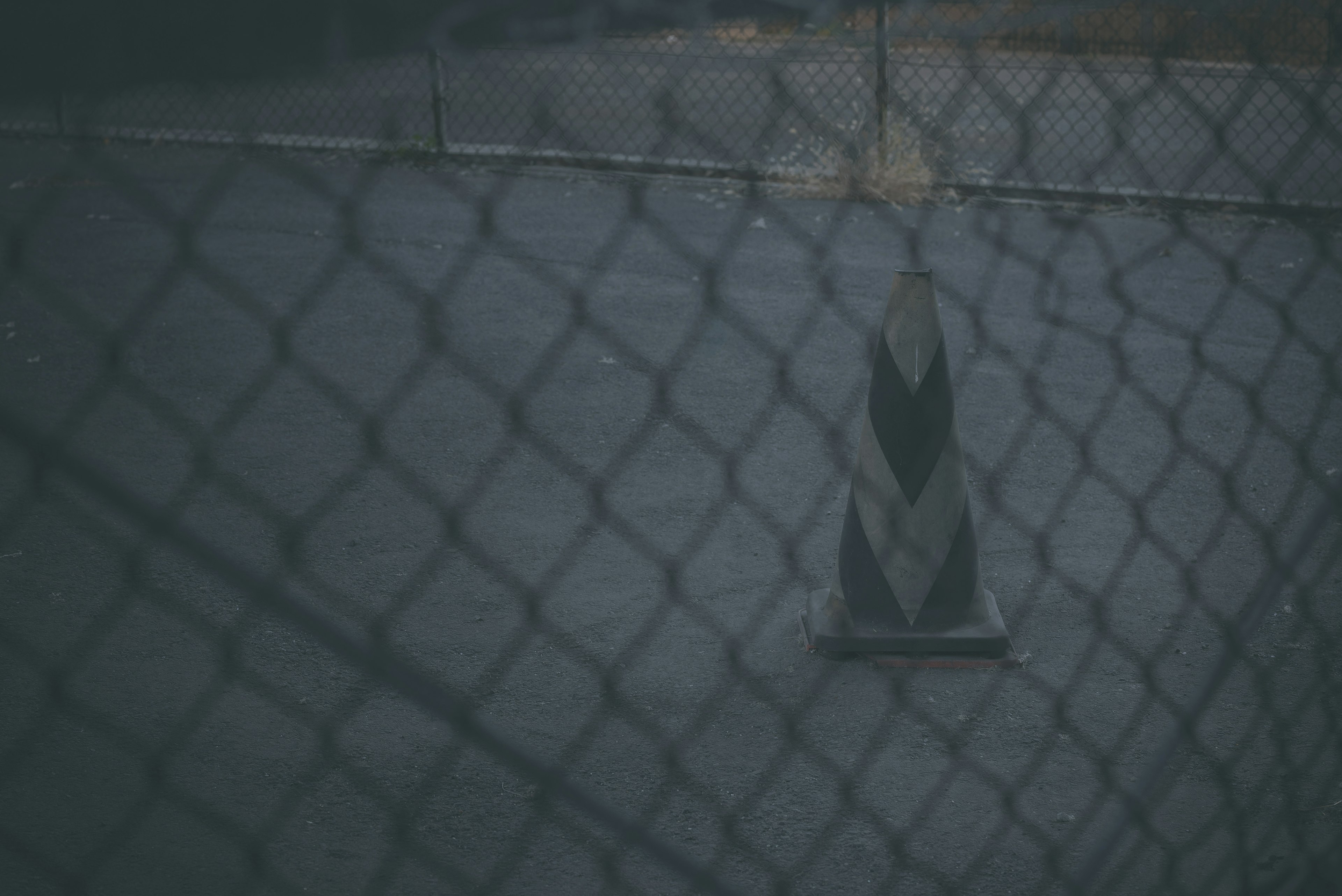 A view of a dark paved surface with a black and white cone in the center seen through a fence