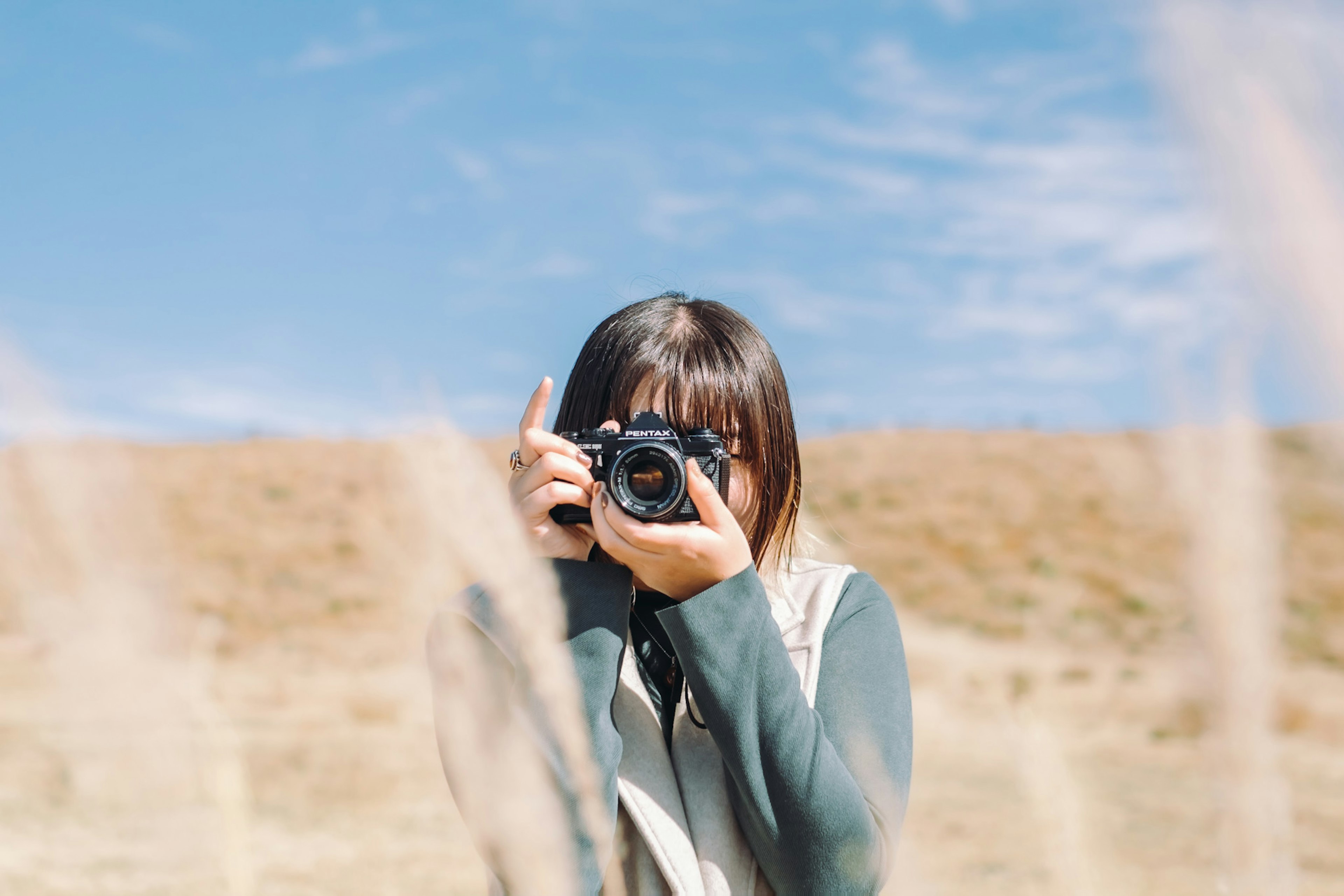 Una mujer sosteniendo una cámara tomando una foto bajo un cielo azul con hierba en primer plano