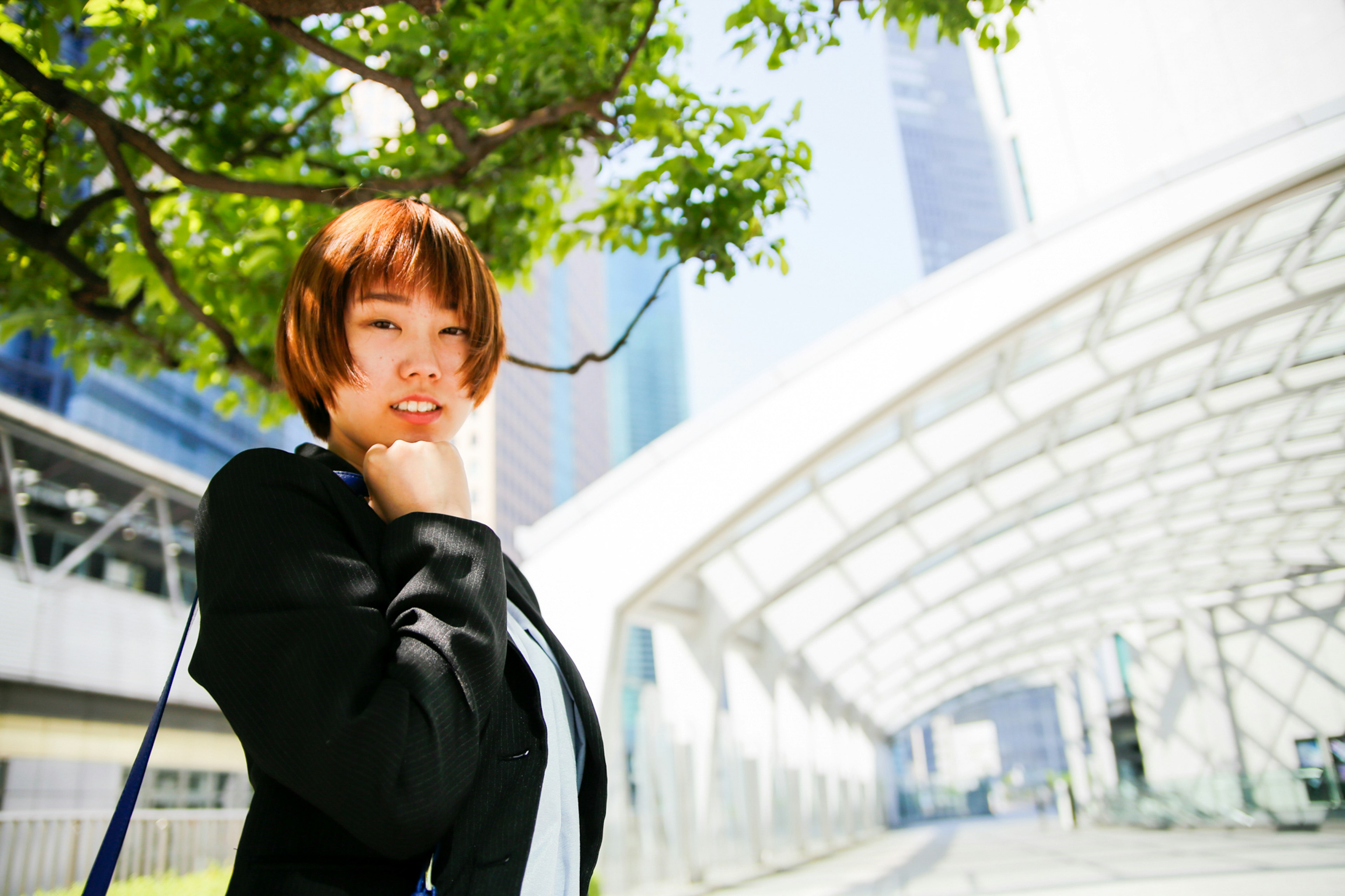 A woman in a business suit smiling in an urban setting with modern buildings and green trees in the background