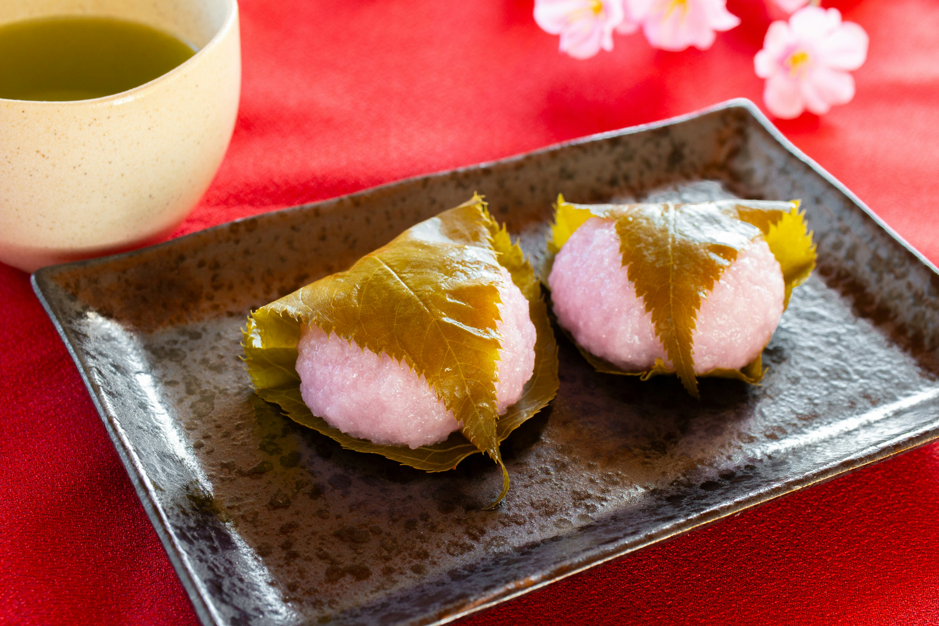 Zwei traditionelle japanische Süßigkeiten auf einem dunklen Teller mit Sakura-Blüten und grünem Tee im Hintergrund