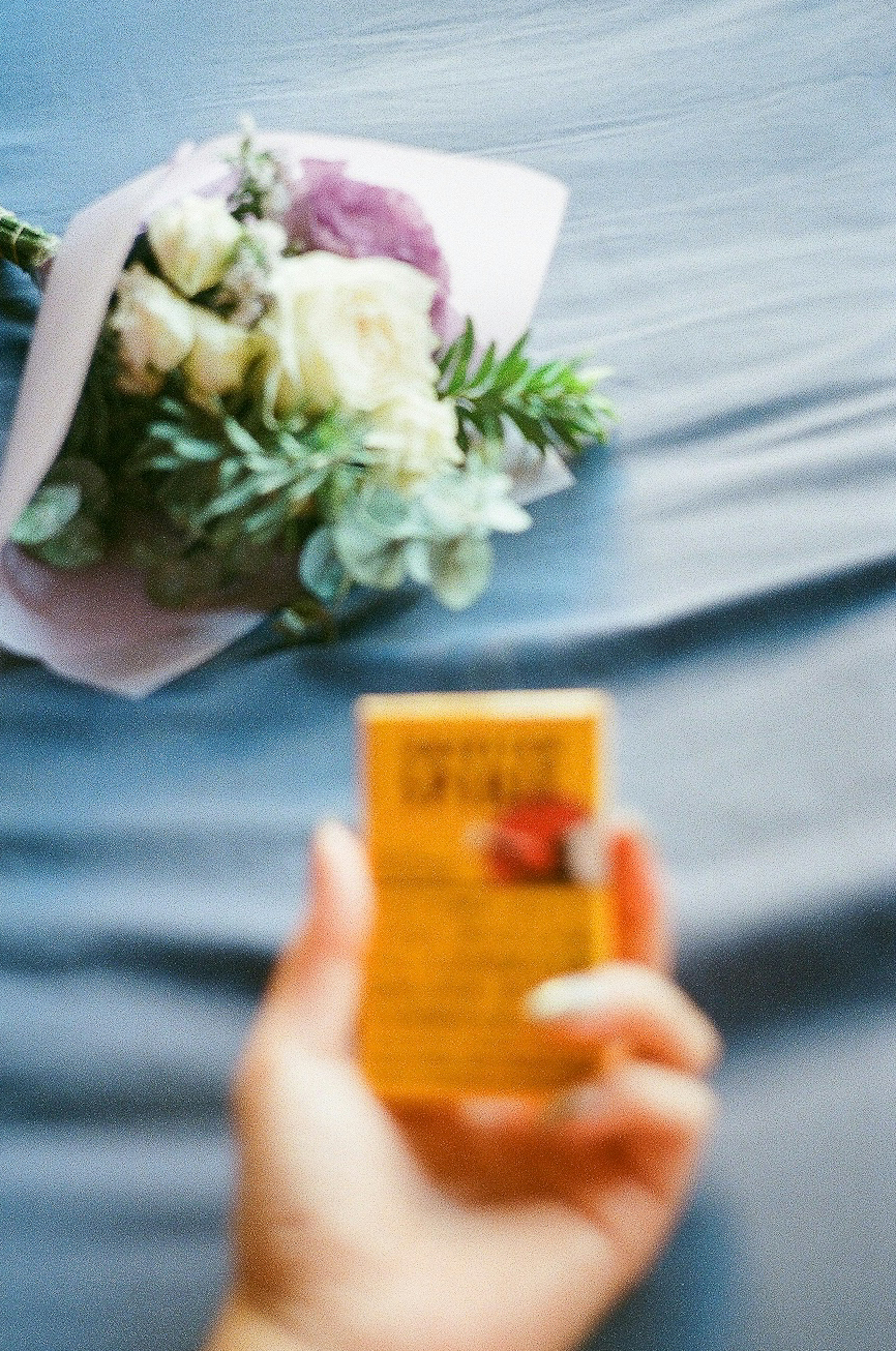 Una mano sosteniendo un paquete amarillo con un ramo de flores al fondo