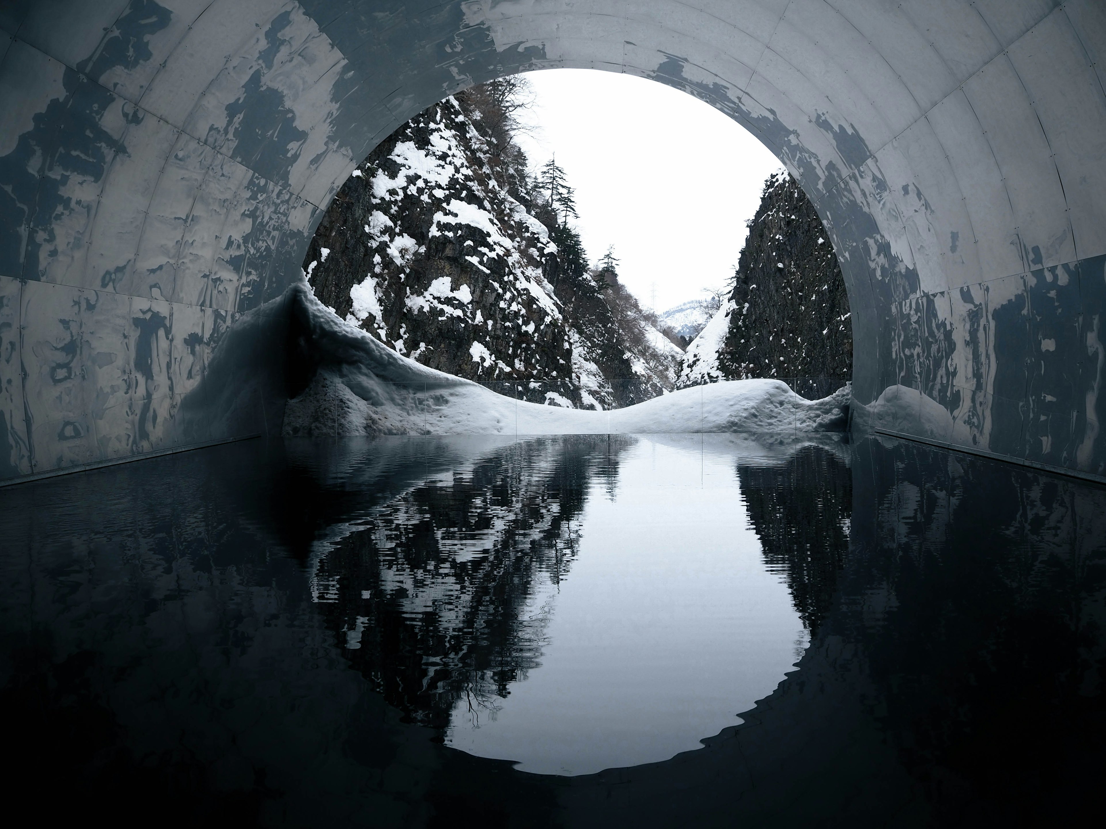 Interior of a concrete pipe showing snow-covered mountains and a calm water surface