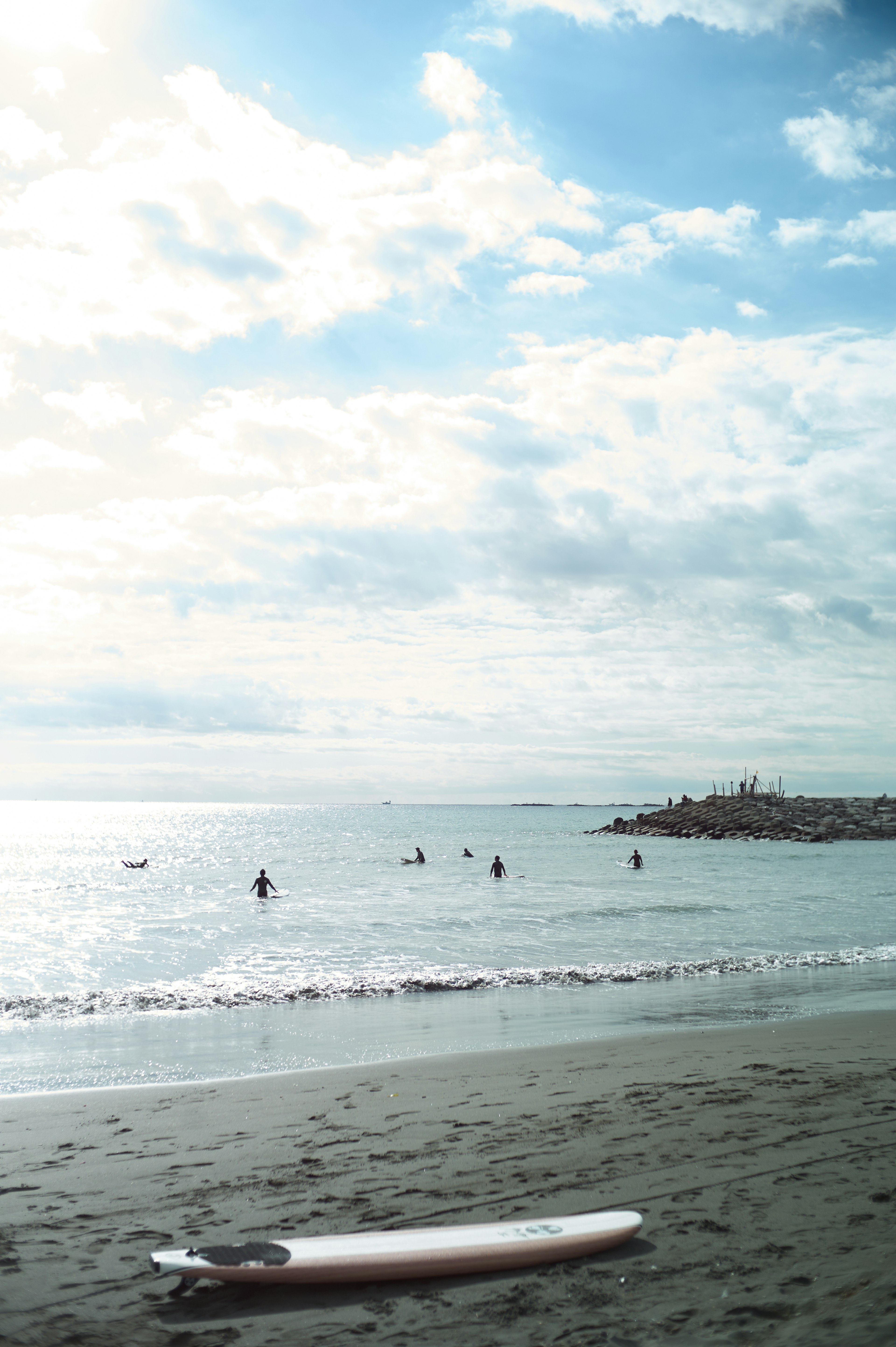 Scena di spiaggia con una tavola da surf sulla sabbia persone che nuotano nel mare sotto un cielo blu con nuvole