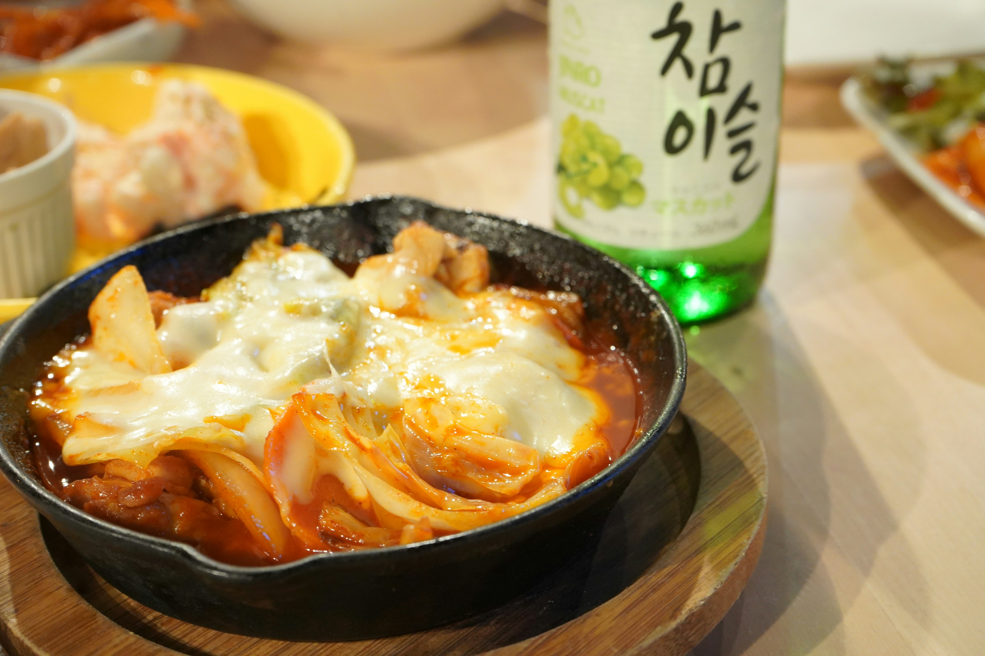 Cheese-topped kimchi stew in a cast iron pot alongside a bottle of Korean drink