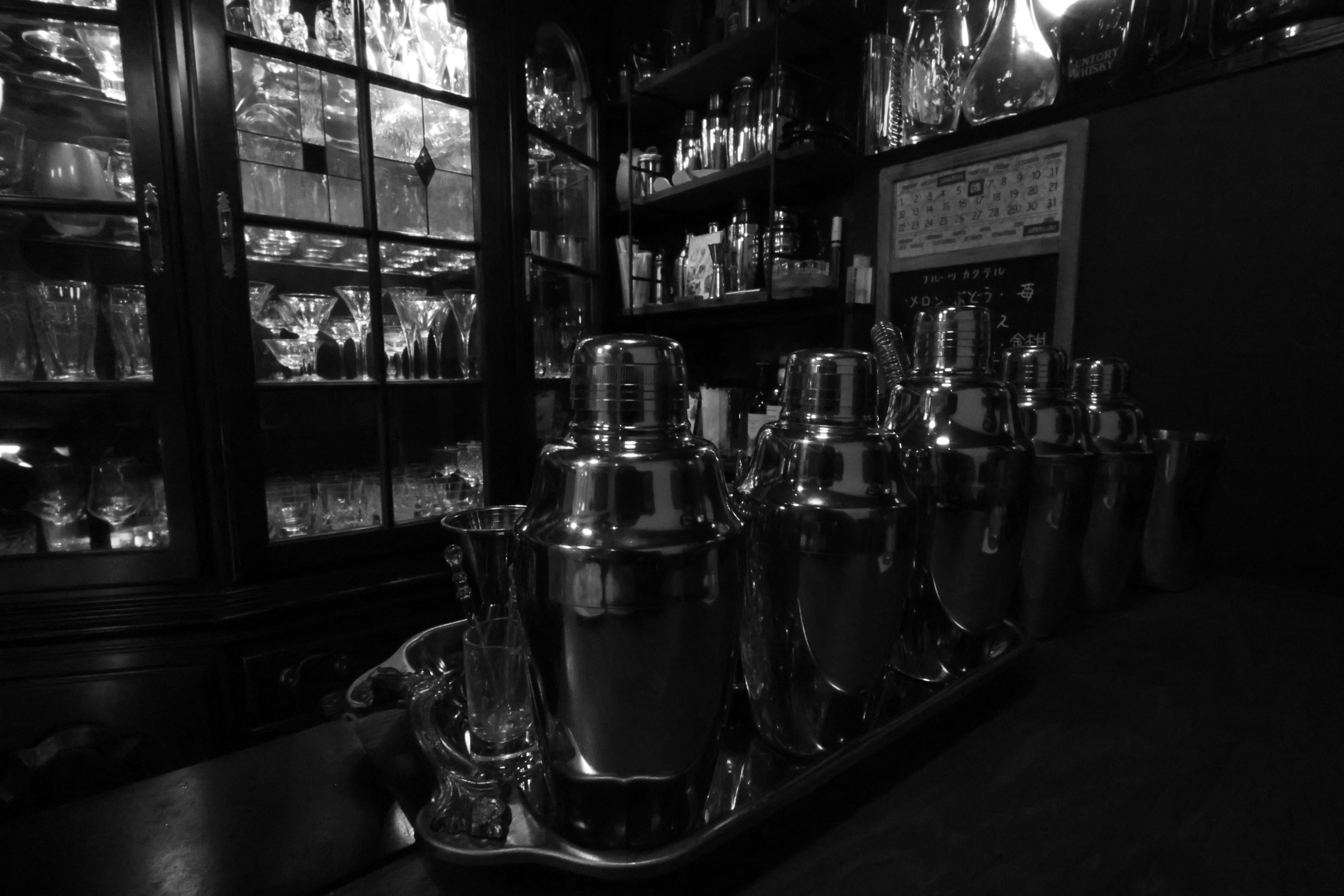 Silver cocktail shakers lined up on a bar counter with a bottle-filled shelf in the background