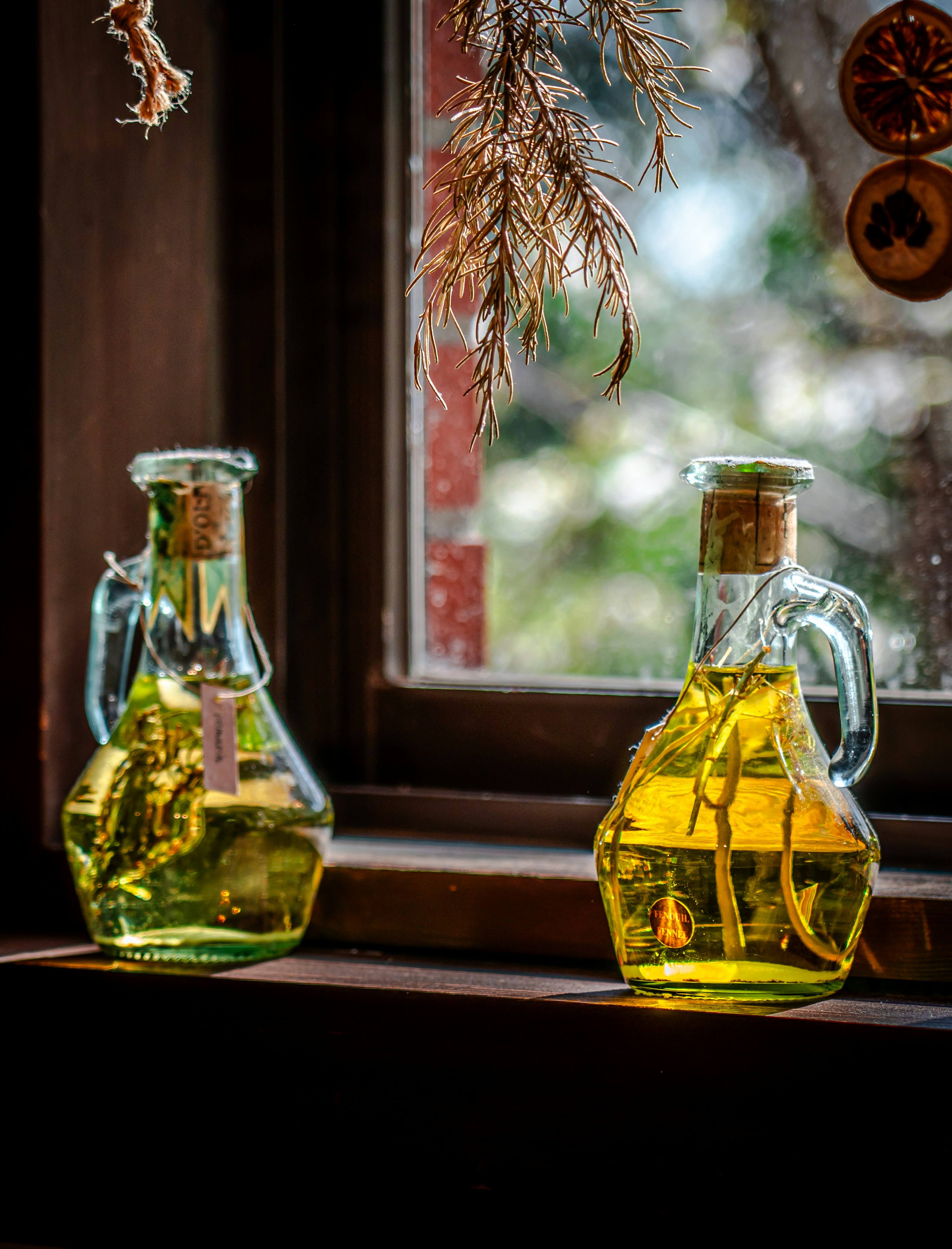 Two bottles of olive oil on a windowsill with dried herbs in the background