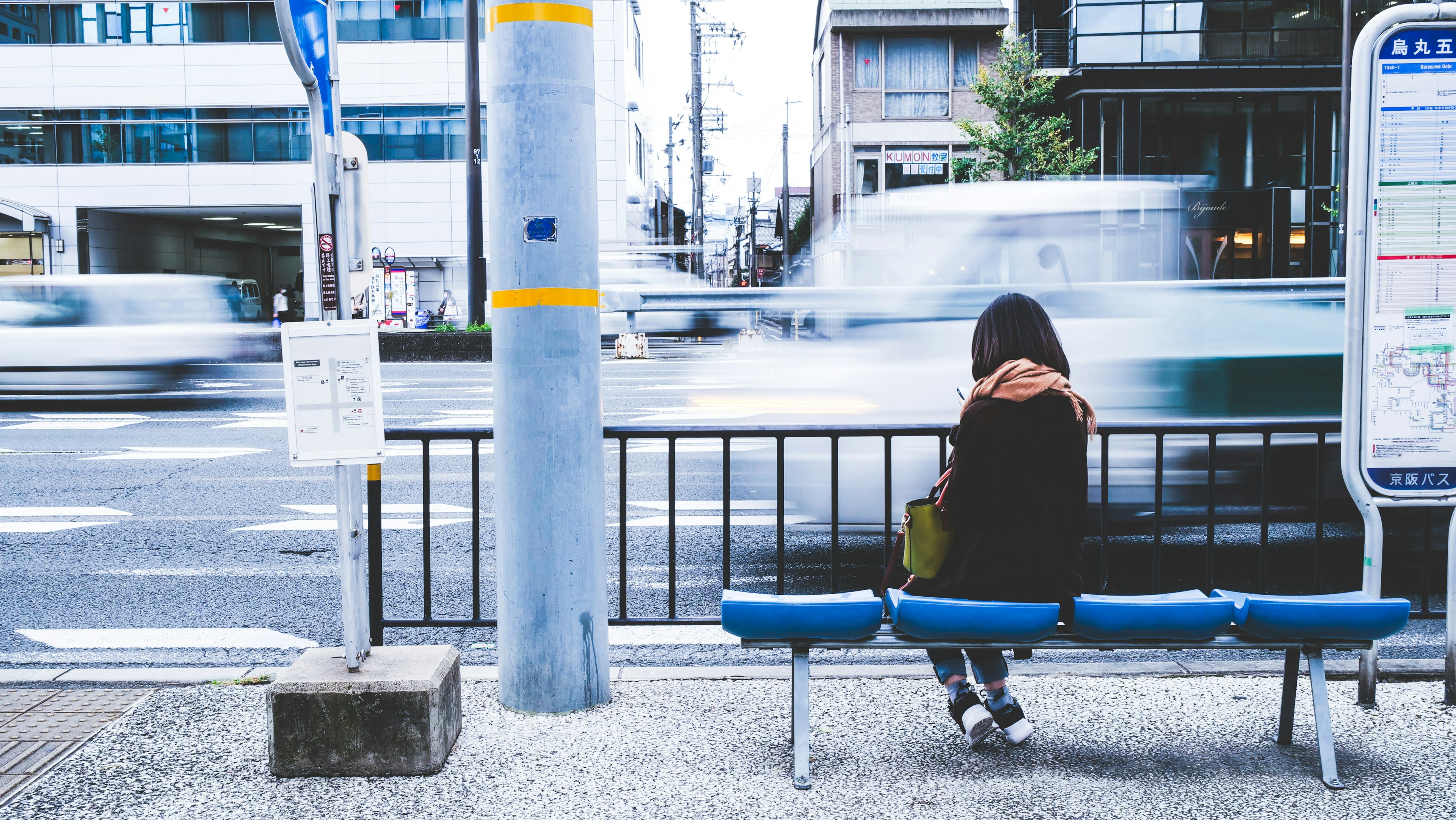 A woman sitting at a bus stop with cars passing by and an urban backdrop