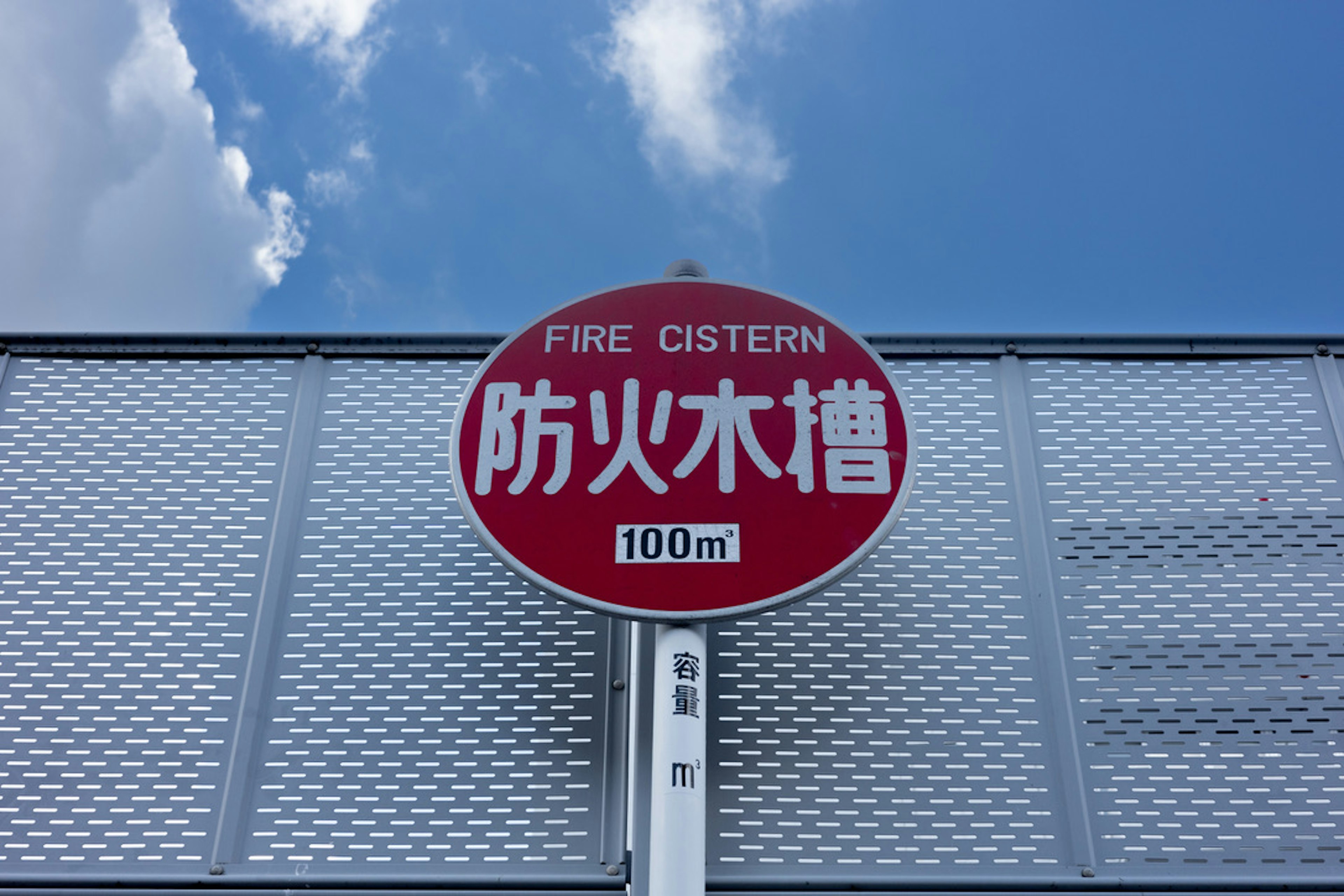 Señal circular roja que muestra el texto 'FIRE CISTERN' y '防火水槽' con un fondo de cielo azul